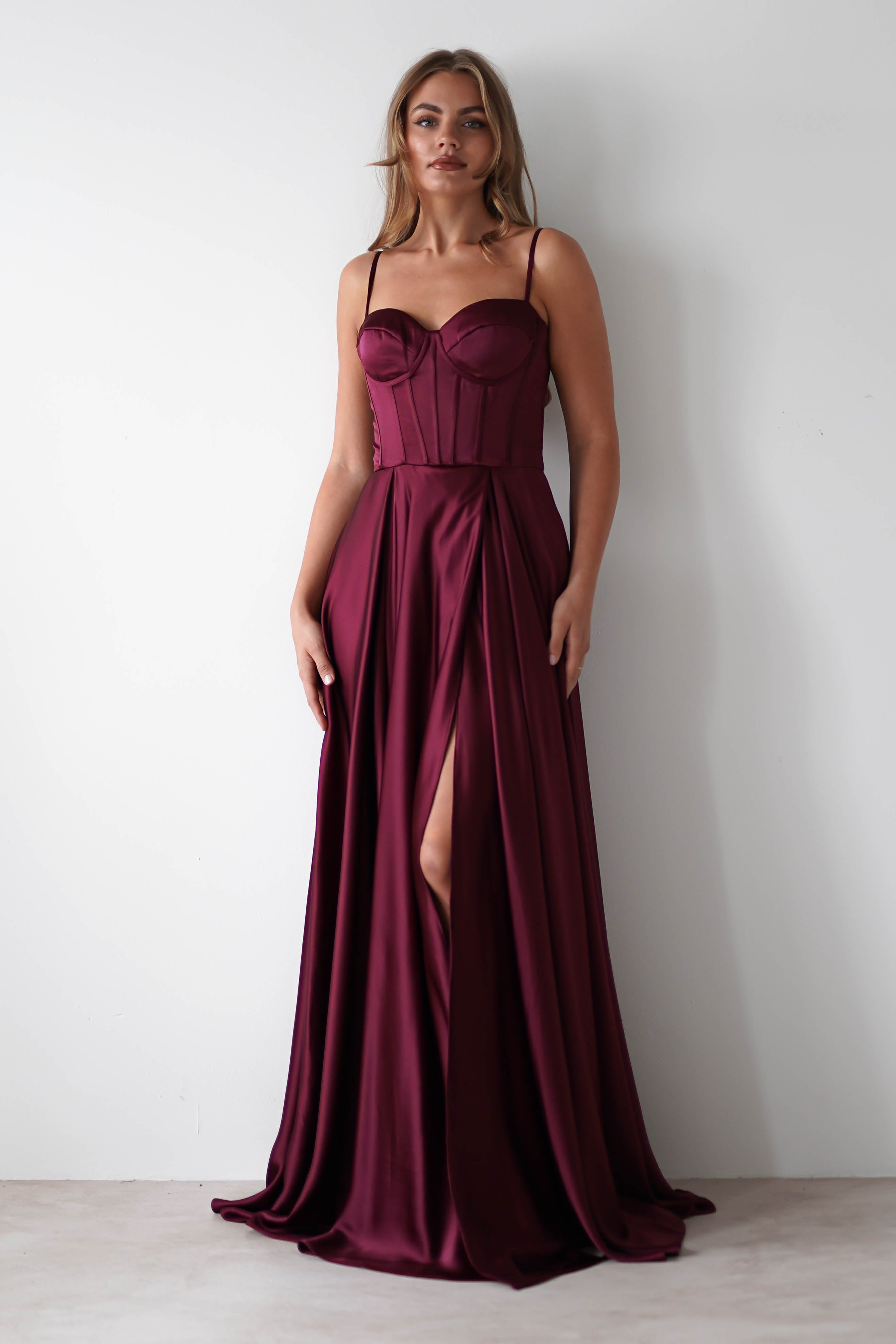 A woman wearing a burgundy satin gown with a fitted bodice, thin straps, lace-up back, and a high front slit stands against a plain white wall—perfect for formal events.
