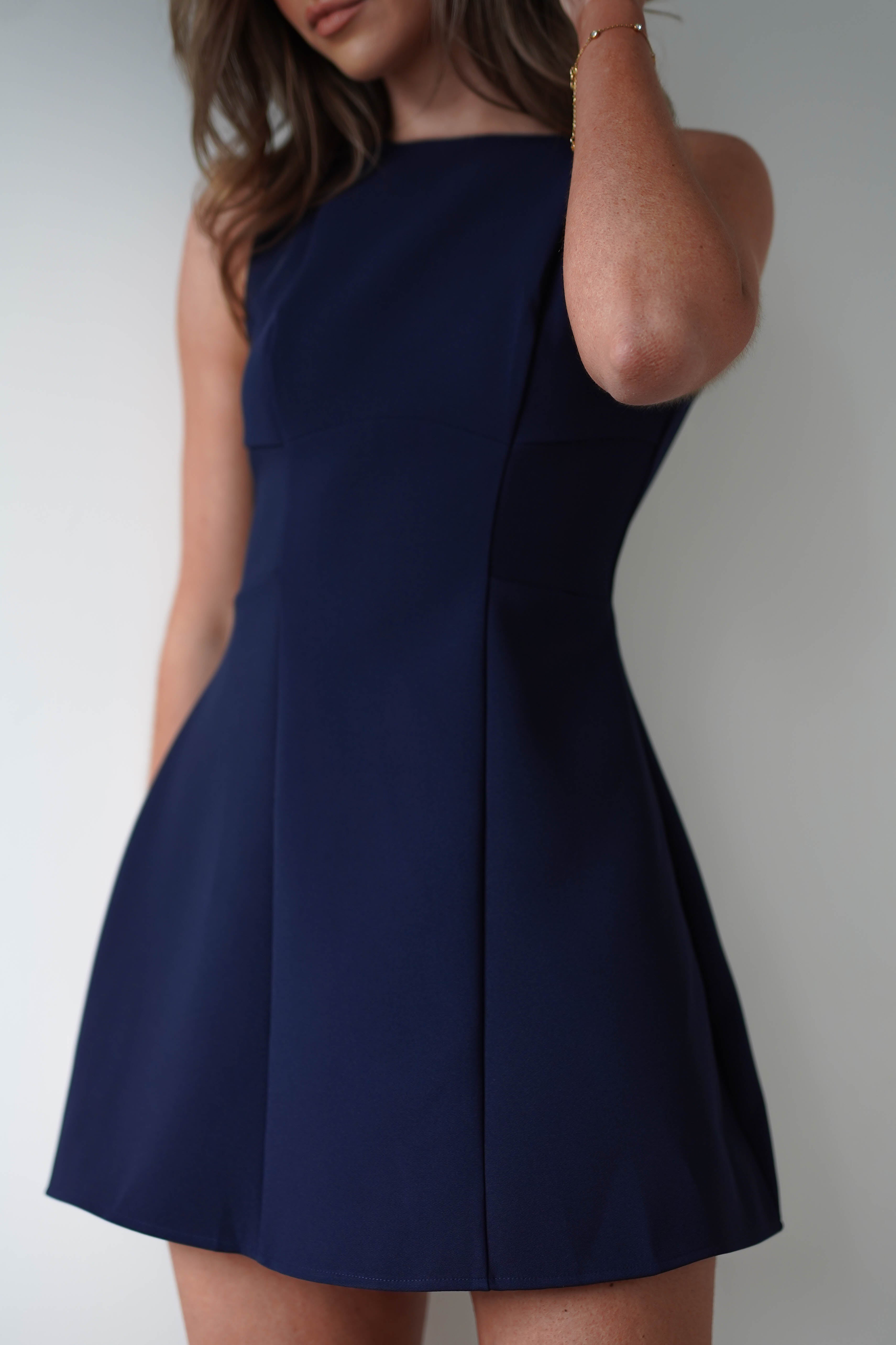 Woman in a sleeveless navy blue mini dress poses against a plain background. Her hand gently touches her hair, and the fully lined dress features an A-line cut that flares out slightly. Perfect for a wedding guest, this look is both elegant and simple.