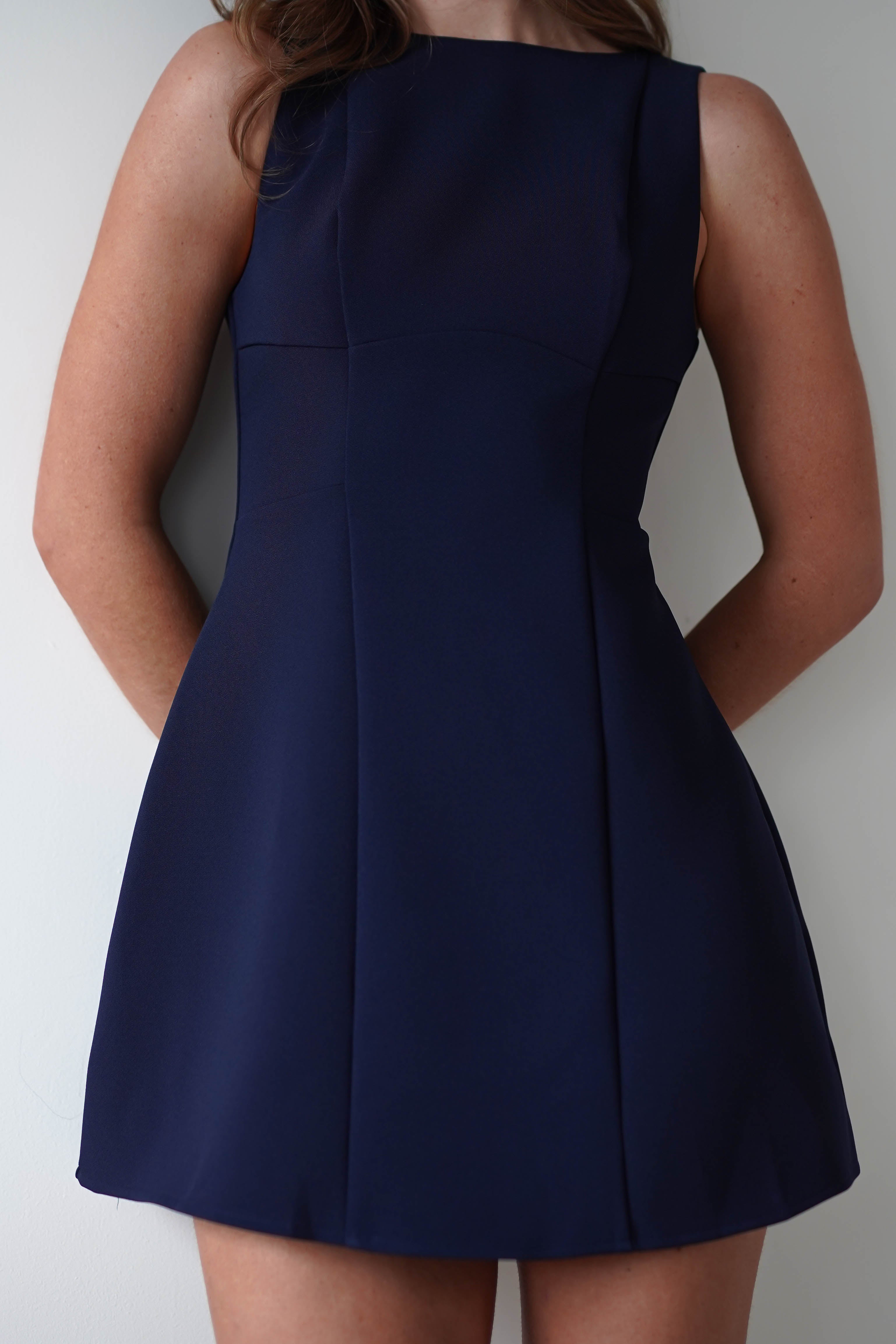 A person in a sleeveless, fitted navy blue mini dress, perfect for a wedding guest, stands against a white wall with arms behind their back. Only the torso and upper legs are visible, showcasing the fully lined elegance of the attire.