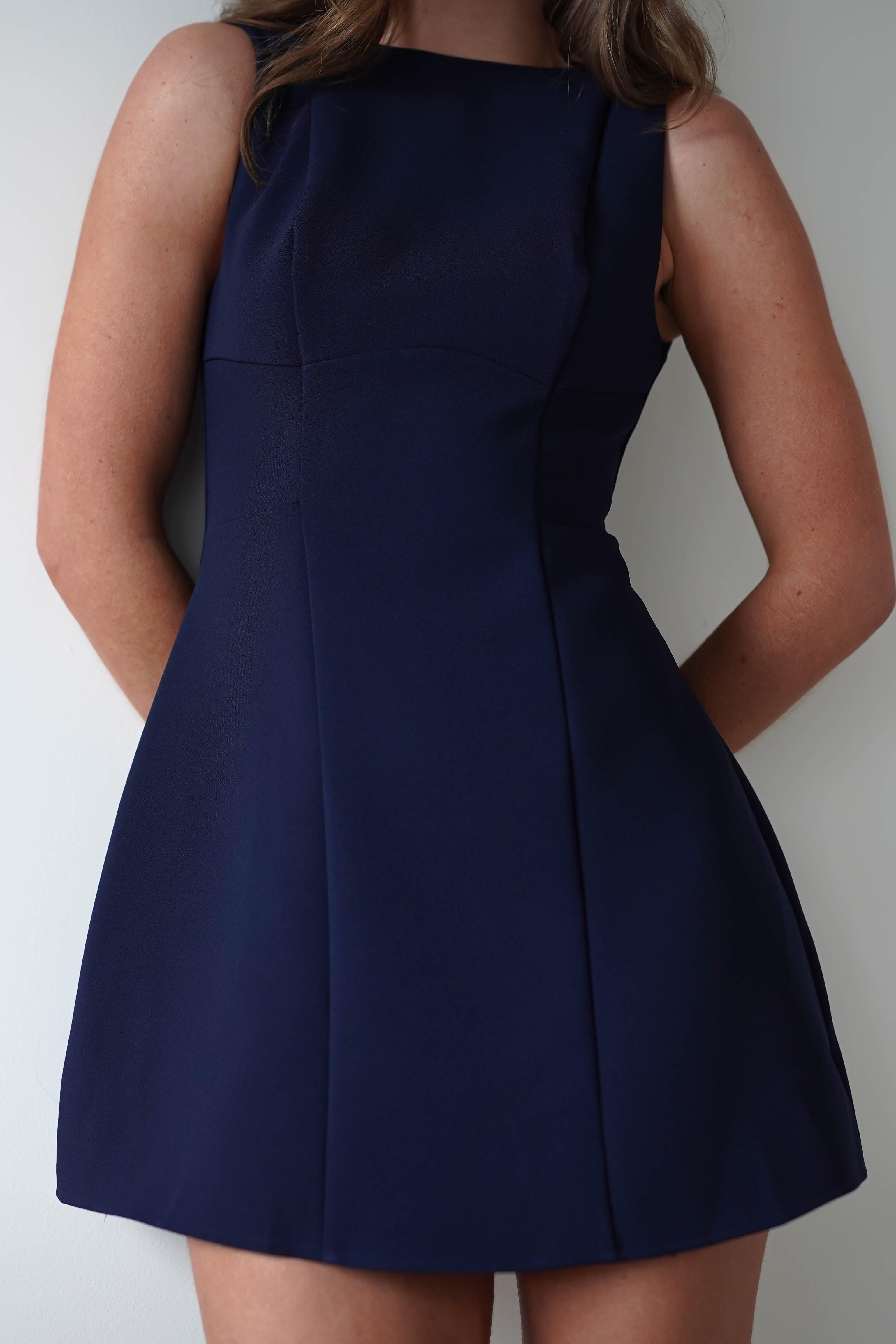 A person wearing a fully lined, sleeveless navy blue mini dress stands poised against a white background. The dress is simple and fitted, highlighting their elegance as a stylish wedding guest. Their arms are positioned behind their back, with the image focusing on the upper bodys refined details.
