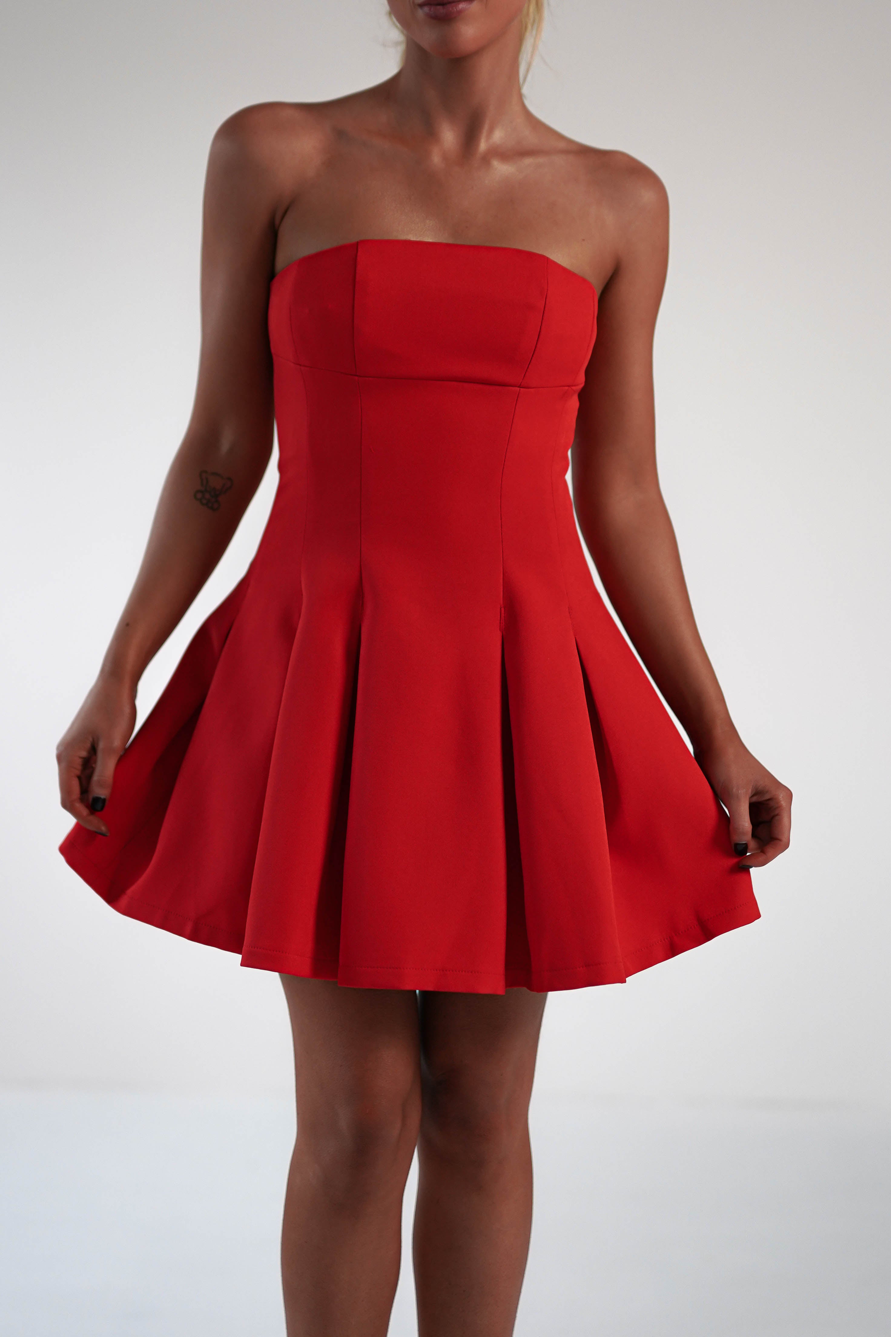 A person in a strapless mini dress, perfect for a wedding guest, holds the sides of their pleated skirt. The fitted bodice complements the vibrant red polyester fabric. A tattoo graces their arm against a plain, light-colored background.