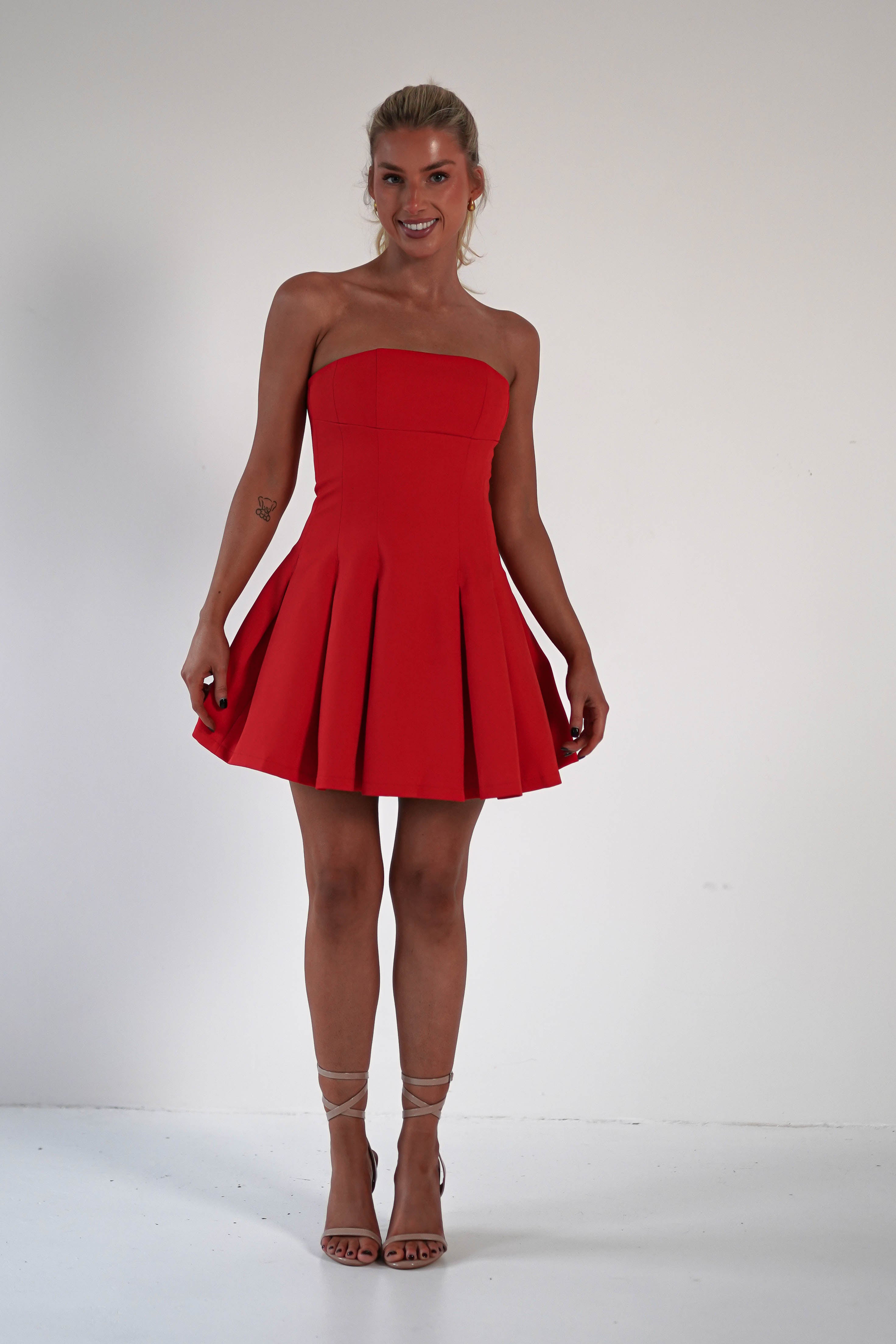 A person stands smiling against a plain background, wearing a strapless red dress with pleats, perfect as a wedding guest dress, paired with beige high-heeled sandals. Their hair is tied back.