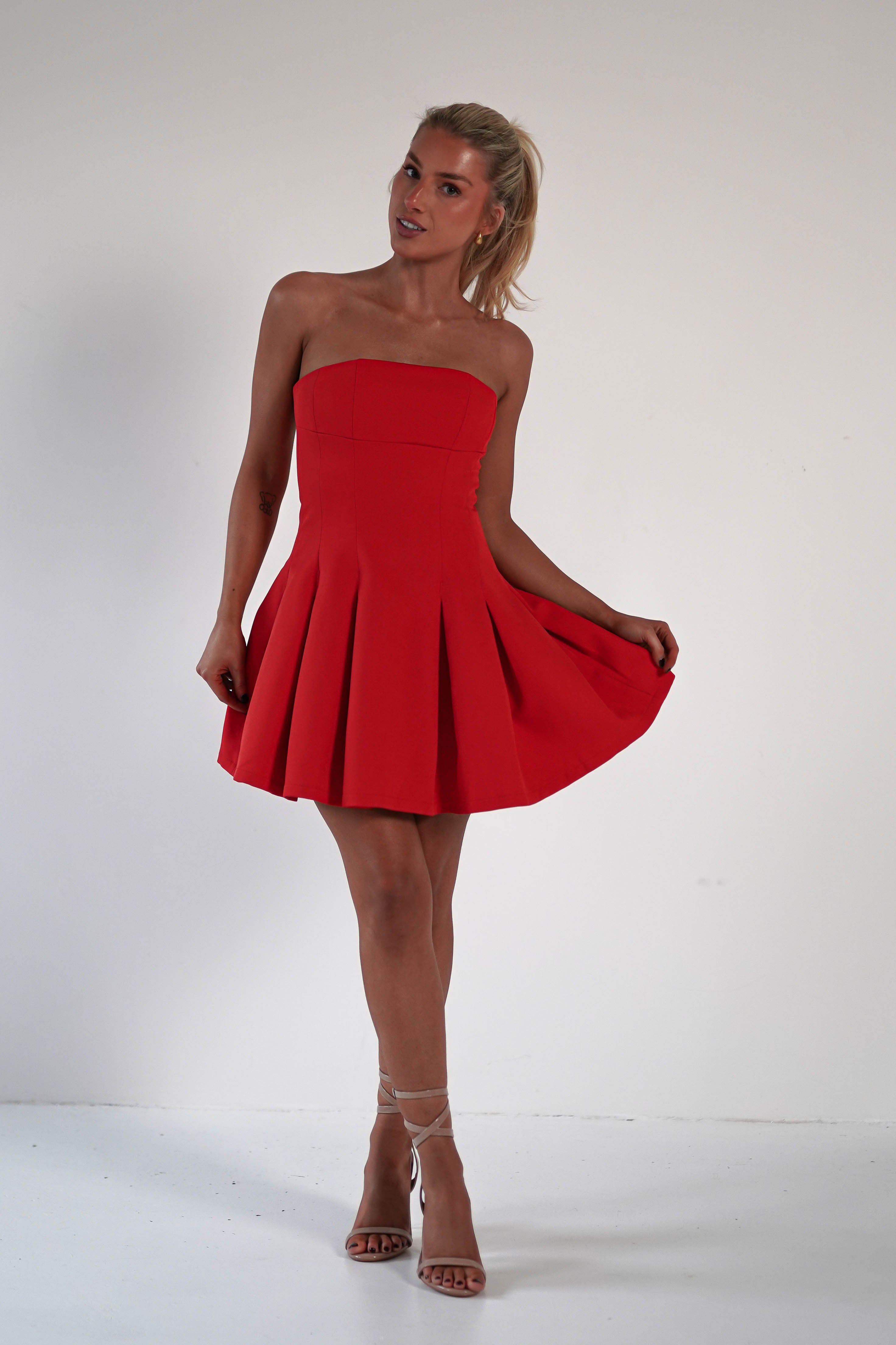 A person in a strapless red wedding guest dress featuring a pleated skirt and light-colored high heels stands against a plain white background. They hold the sides of the polyester dress, slightly lifting it. Hair is styled in a ponytail.