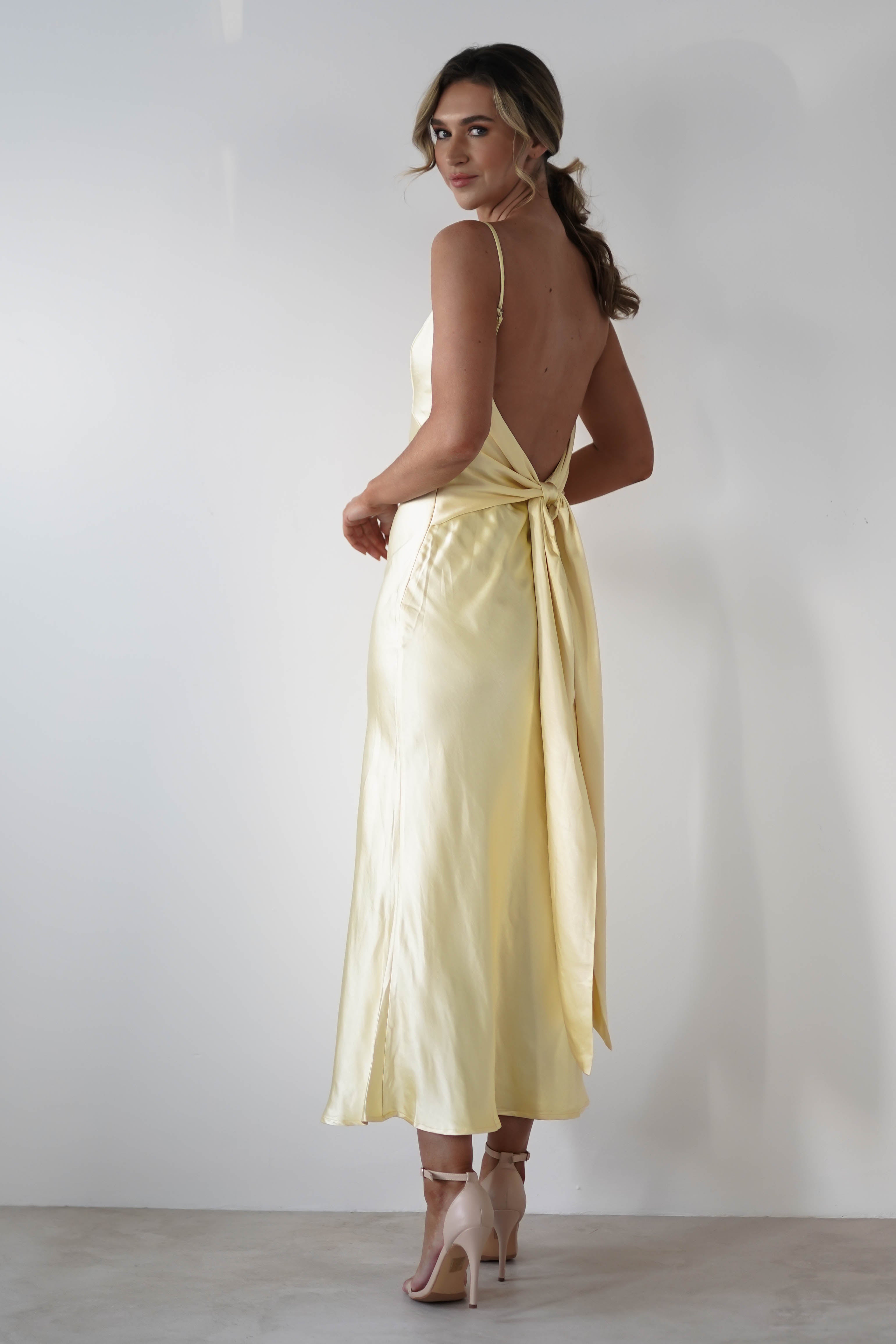 A woman in a sleeveless, backless pale yellow satin knit midi dress with a hidden side zipper stands against a plain white wall, facing away and looking back over her shoulder. Perfect for bridesmaids, she wears high-heeled shoes and her hair is in loose waves.