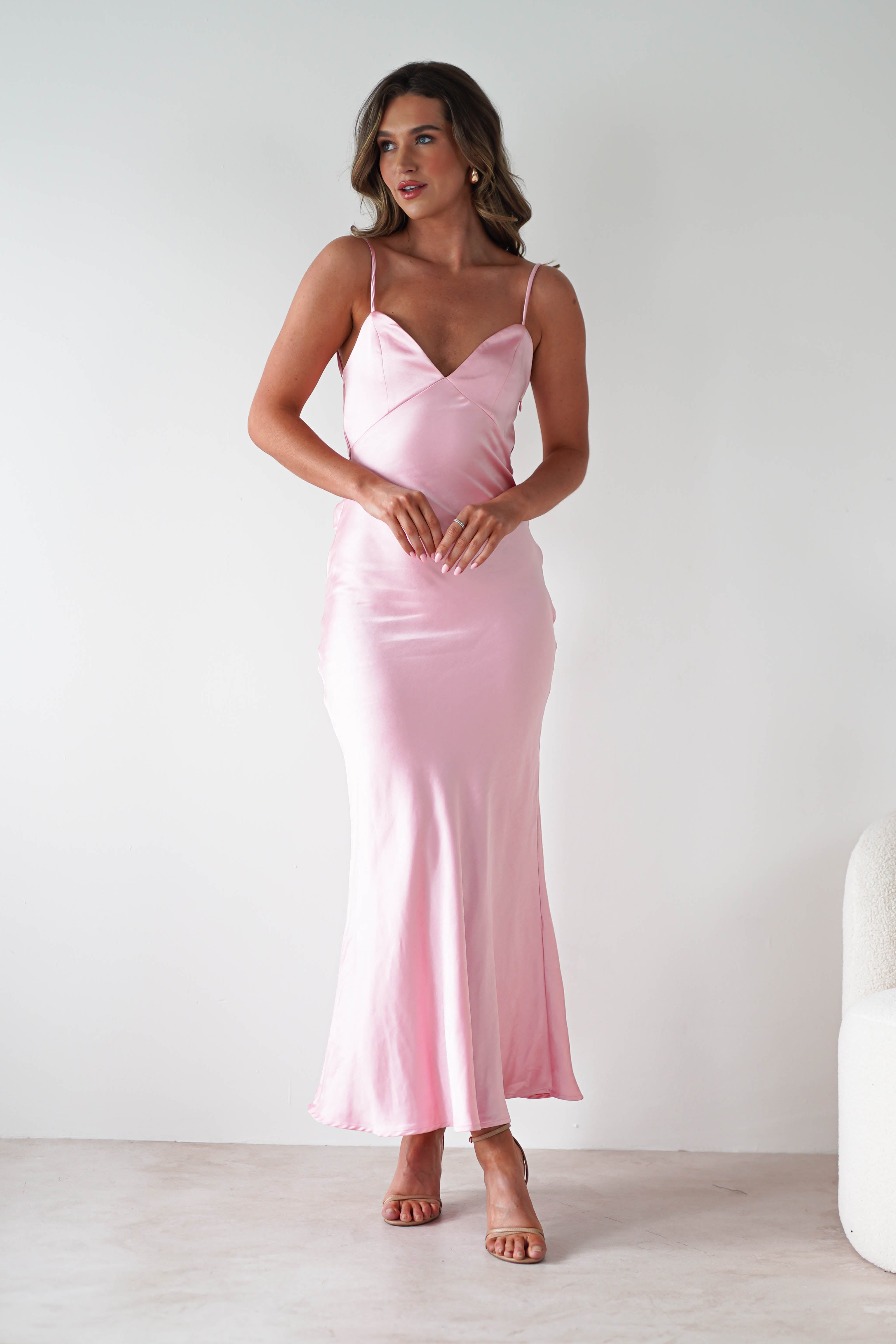 A woman stands against a plain white background, wearing a silky light pink satin dress with thin straps and a V-neckline—a perfect choice for bridesmaids. She has wavy hair and her hands are gently clasped in front of her.