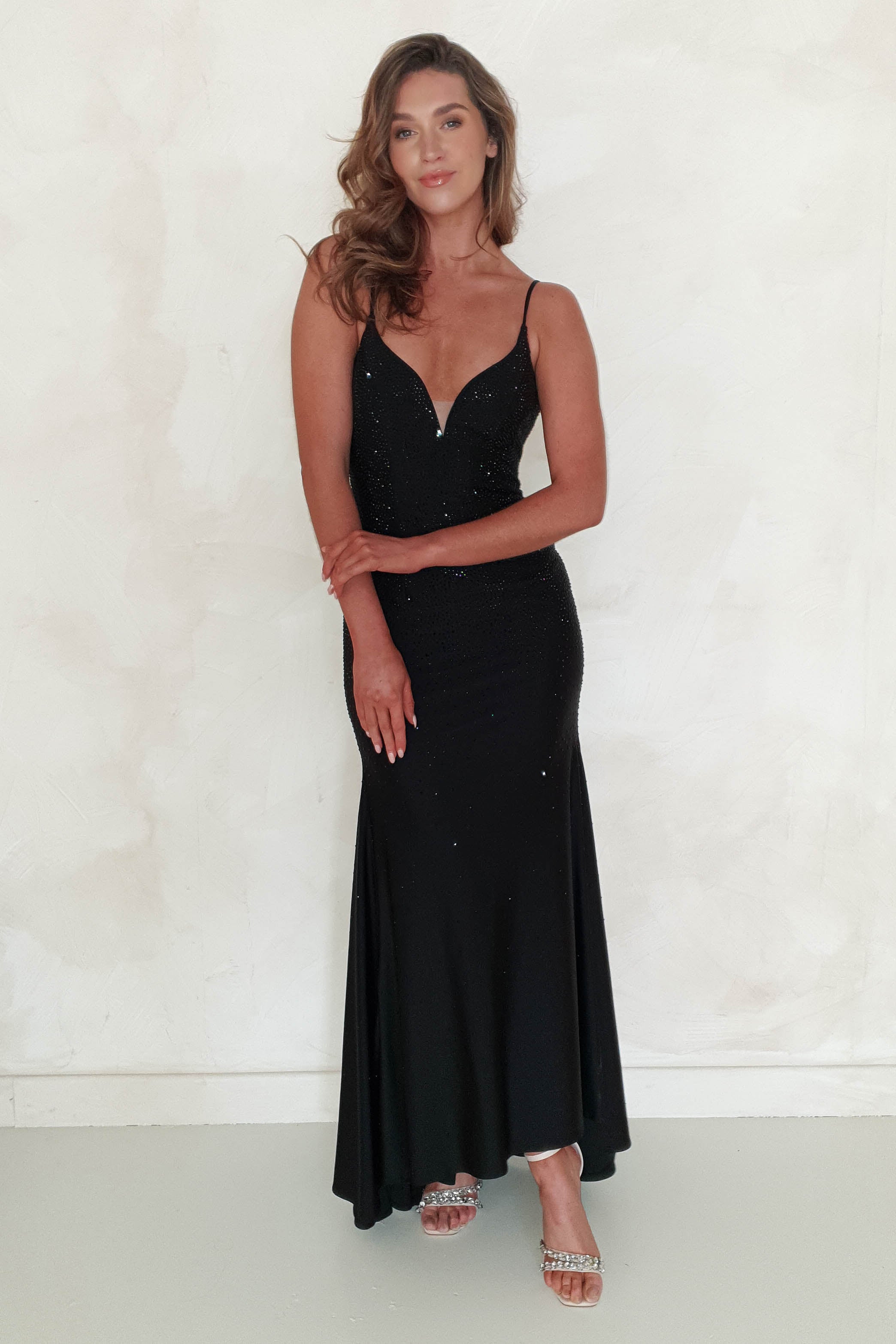 A woman with wavy brown hair stands against a light-colored wall, wearing a long, fitted diamante gown with thin straps and sparkly details—perfect for formal occasions. She pairs her look with silver open-toe heels and smiles softly, hands clasped in front.