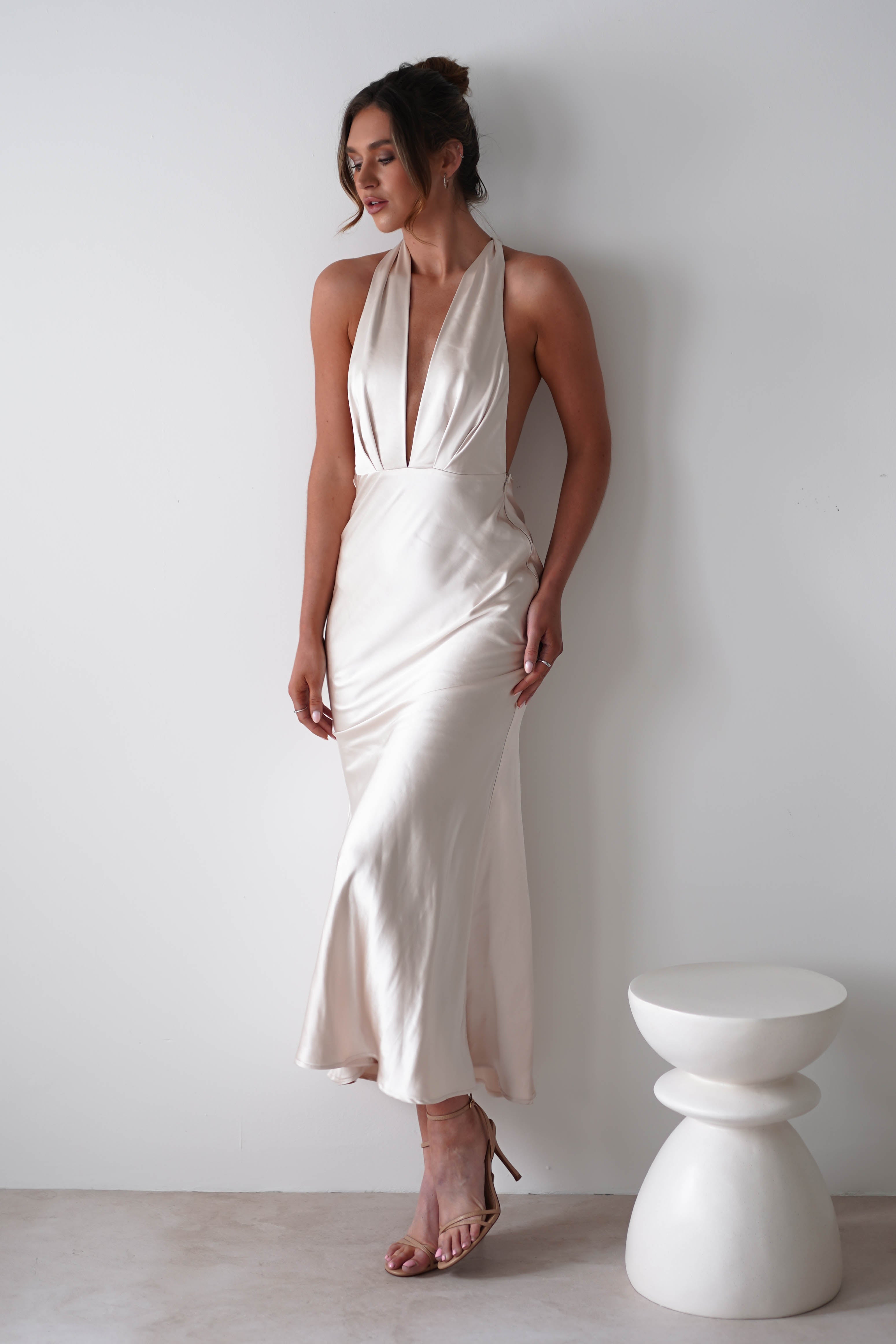 A woman stands against a white wall wearing a soft satin material, cream-colored halter-neck dress with a deep V neckline and open sides—perfect for rehearsal dinners. She has her hair up, strappy heels on, with a modern white stool beside her.