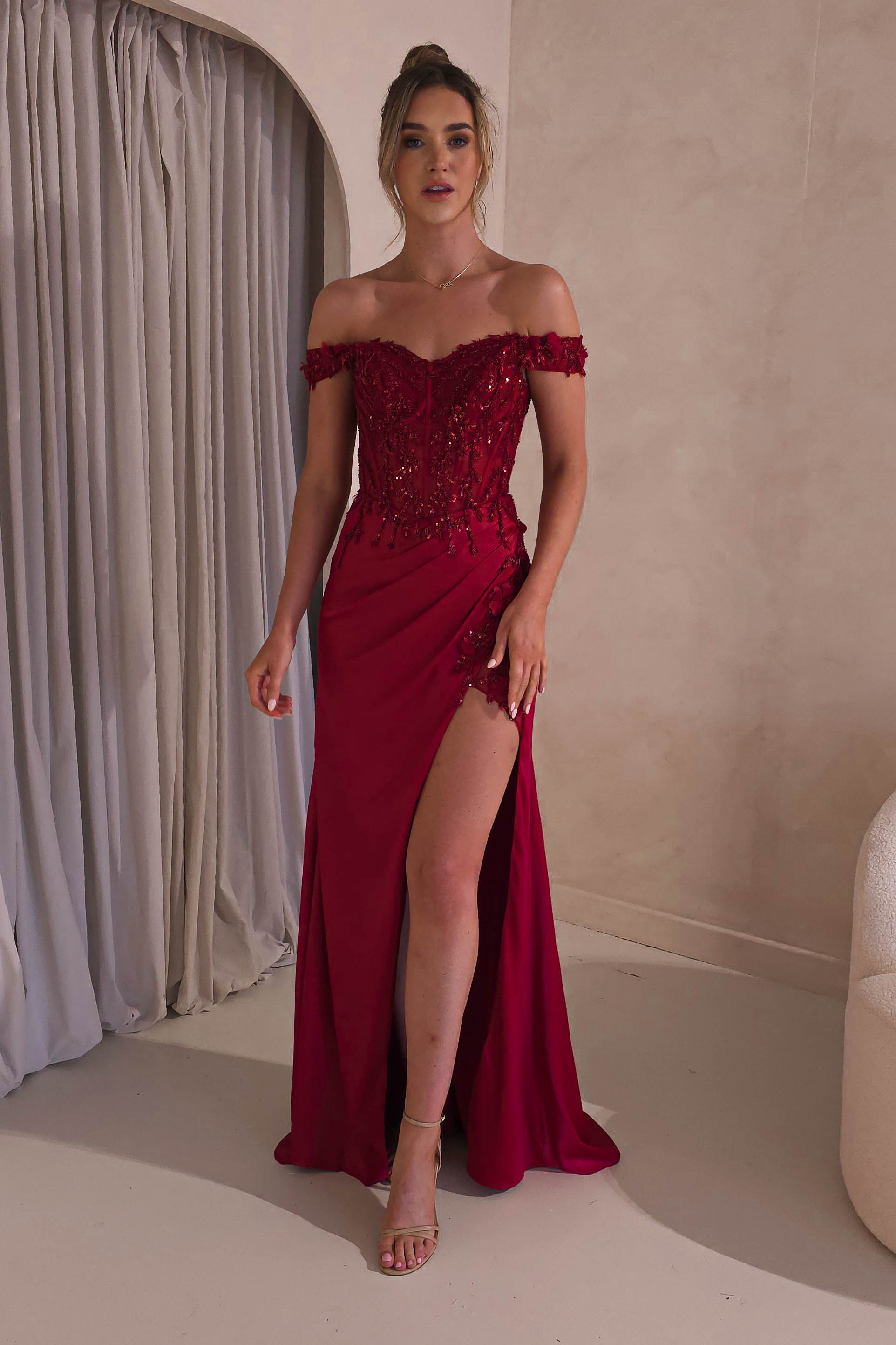 A woman stands in a room wearing a satin gown with intricate embellishment details. The off-shoulder design features a sweetheart neckline and a thigh-high slit. Her hair is styled up, and she poses elegantly near a curtain.