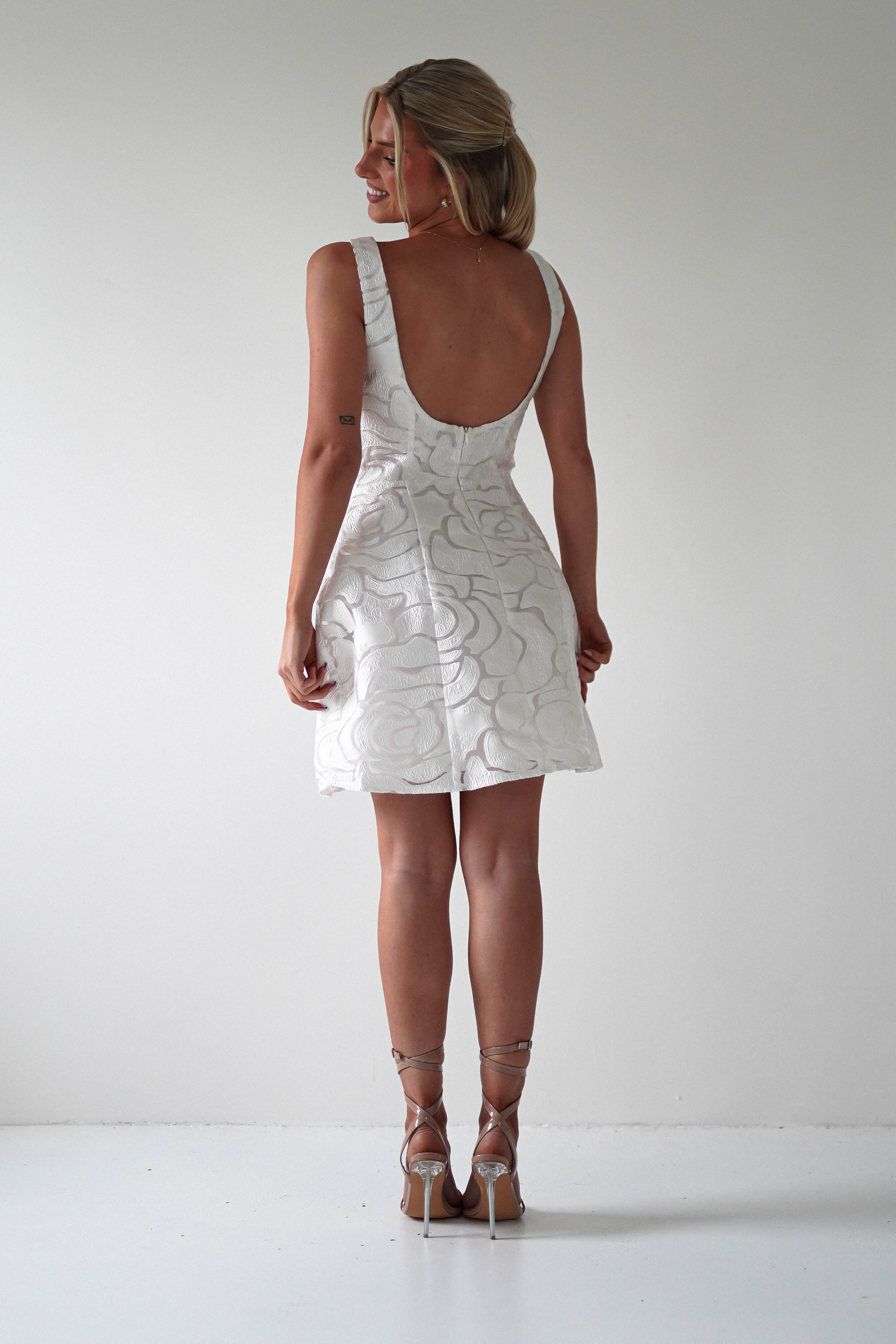 A woman stands smiling in an embroidered mesh dress with a floral pattern and low back, perfect for a wedding guest. She wears strappy high heels, her hair stylishly up as she poses against a plain white background, looking over her shoulder.