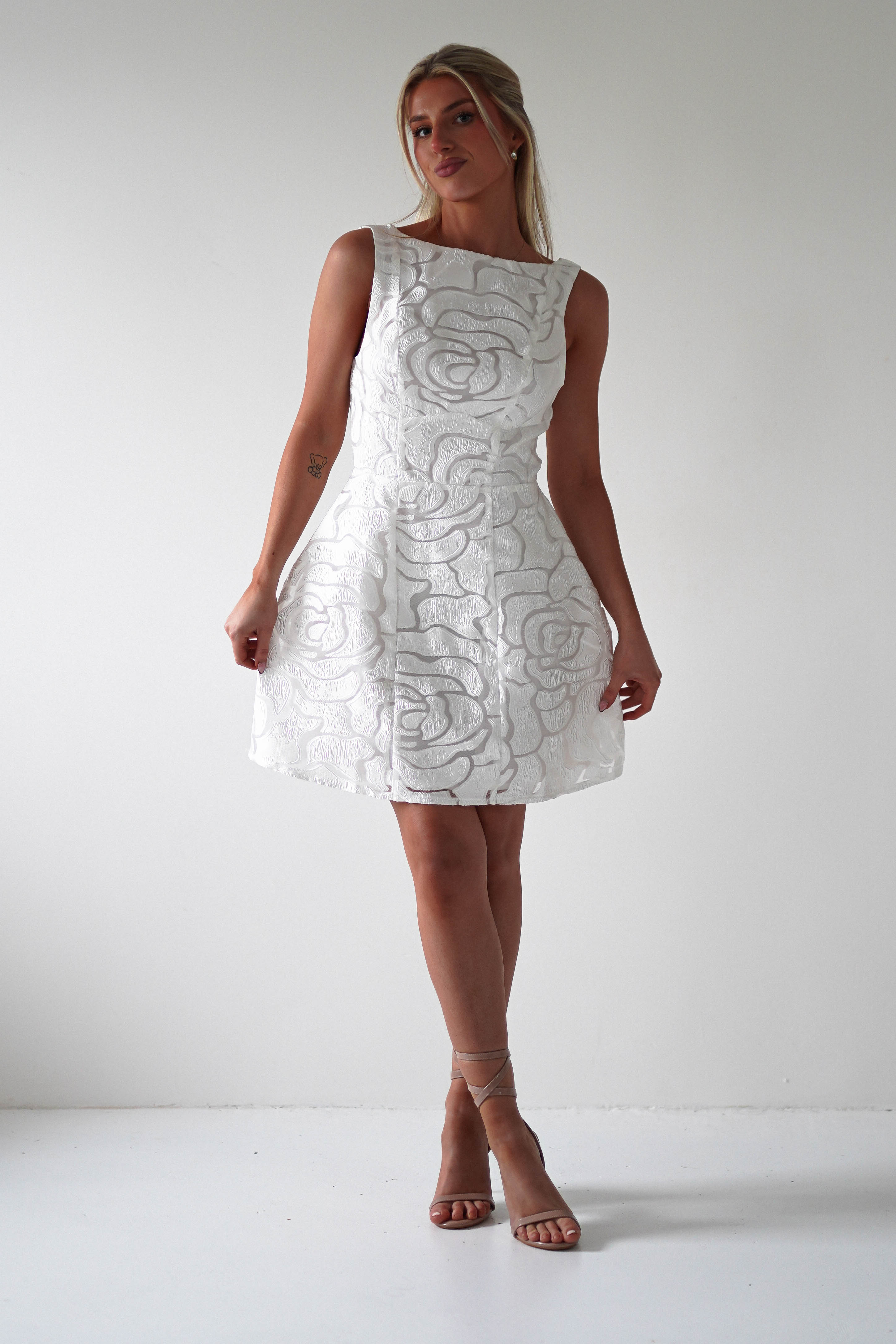 A person in an elegant sleeveless embroidered mesh dress with floral patterns stands against a plain white background. Their strappy sandals complement the look, while their hair is pulled back, creating a chic ensemble perfect for graduations or as a stylish wedding guest outfit.