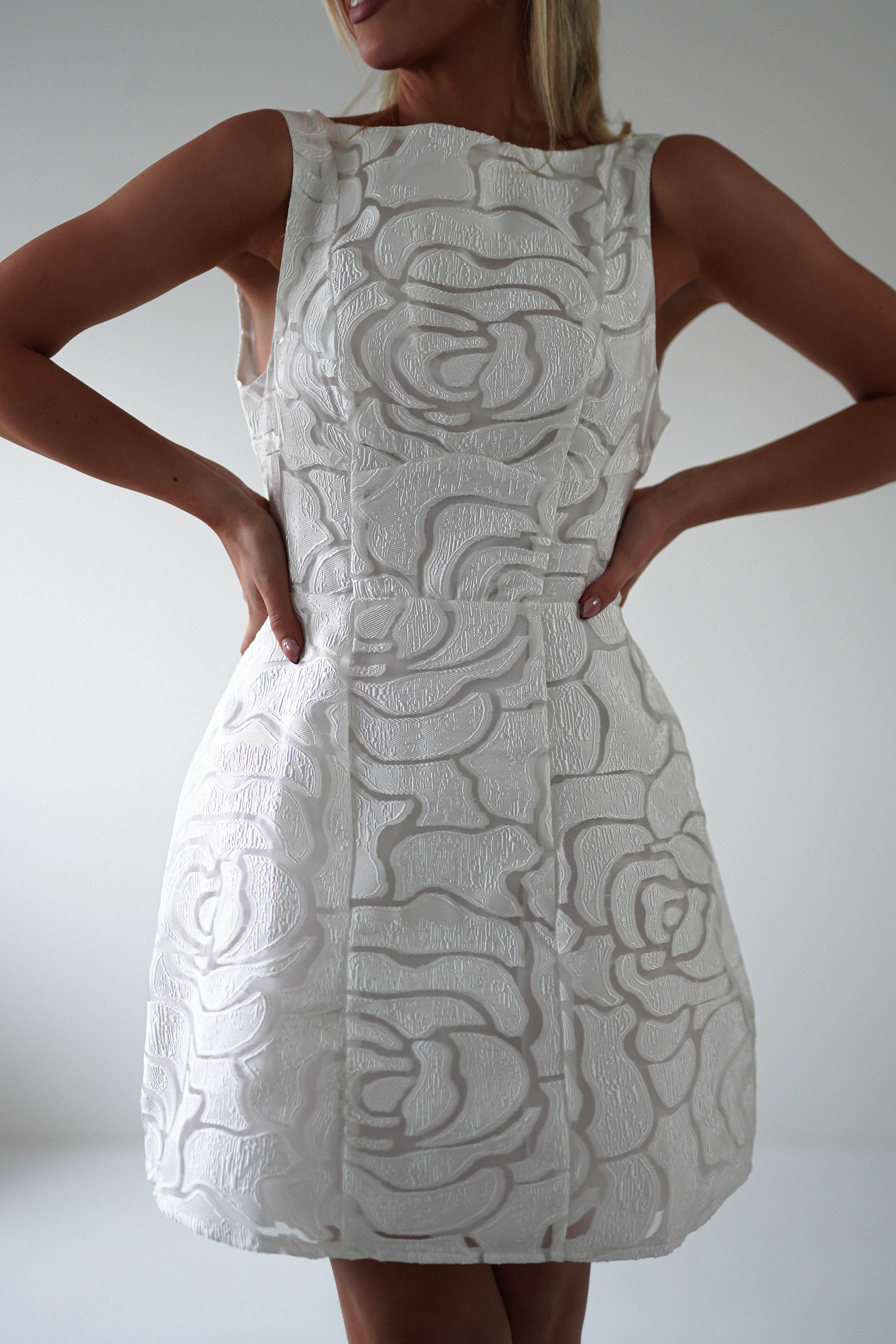 A person wearing an elegant sleeveless white dress with an embroidered mesh floral pattern stands against a plain background, hands confidently on hips, ready to shine at graduations or as a stylish wedding guest.