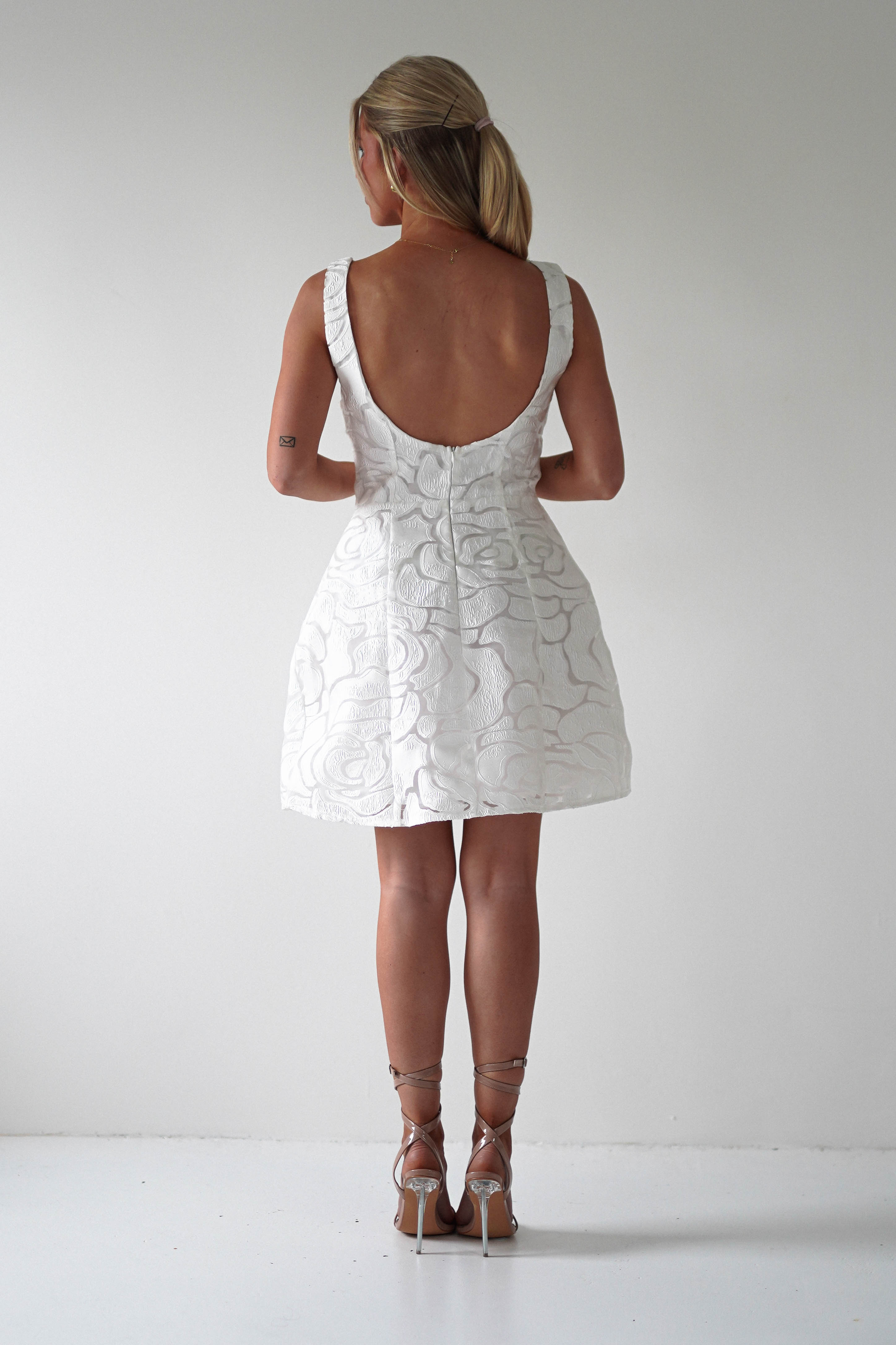 A person stands against a white background wearing an embroidered mesh dress with floral details, perfect for a wedding guest. The sleeveless dress features a low back design. They complement the look with silver high-heeled sandals and have long hair elegantly tied back.
