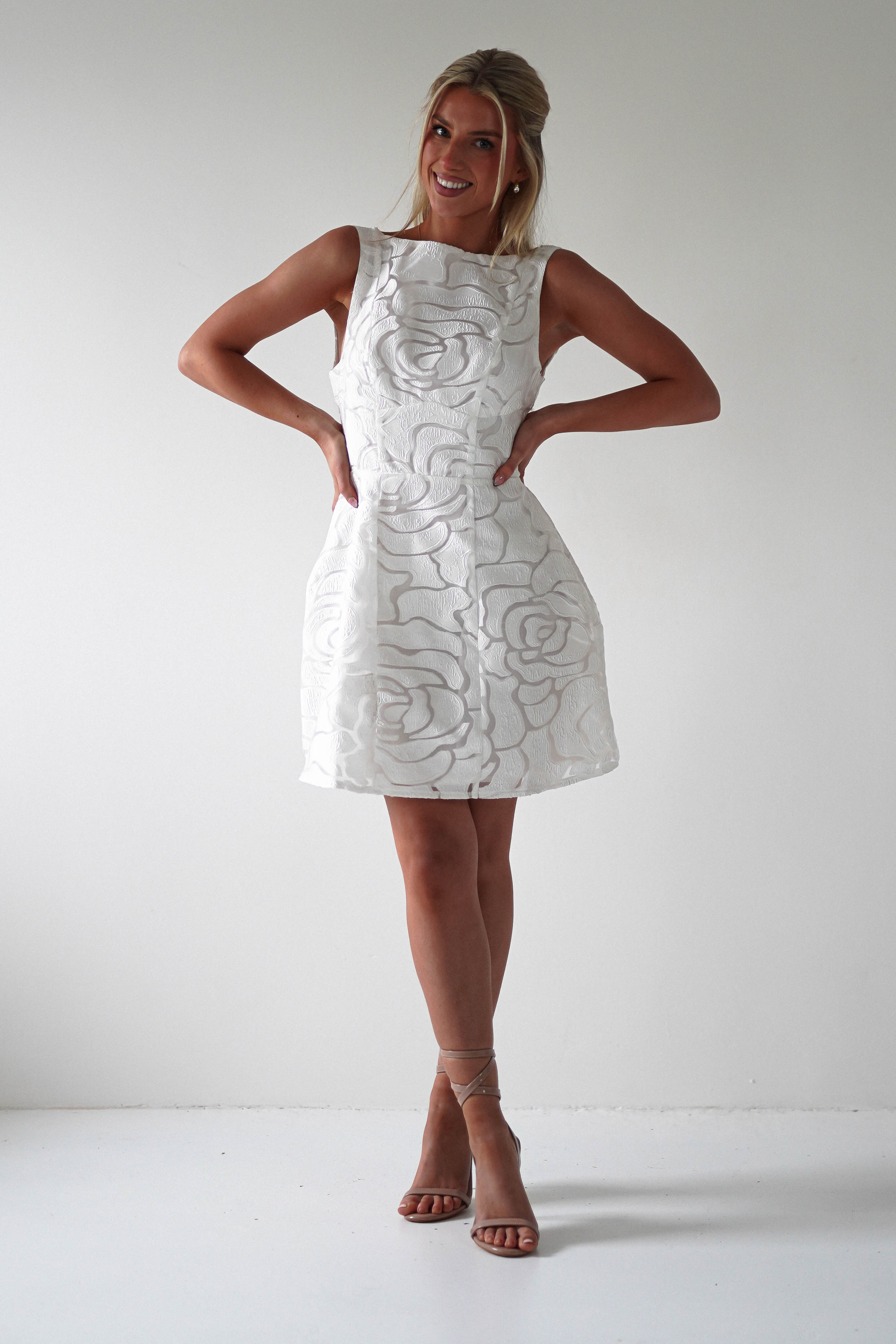 A person in a sleeveless embroidered mesh dress with a floral pattern stands confidently with hands on hips, embodying perfect wedding guest style. They wear strappy sandals and have an elegant updo hairstyle against the plain white wall backdrop.