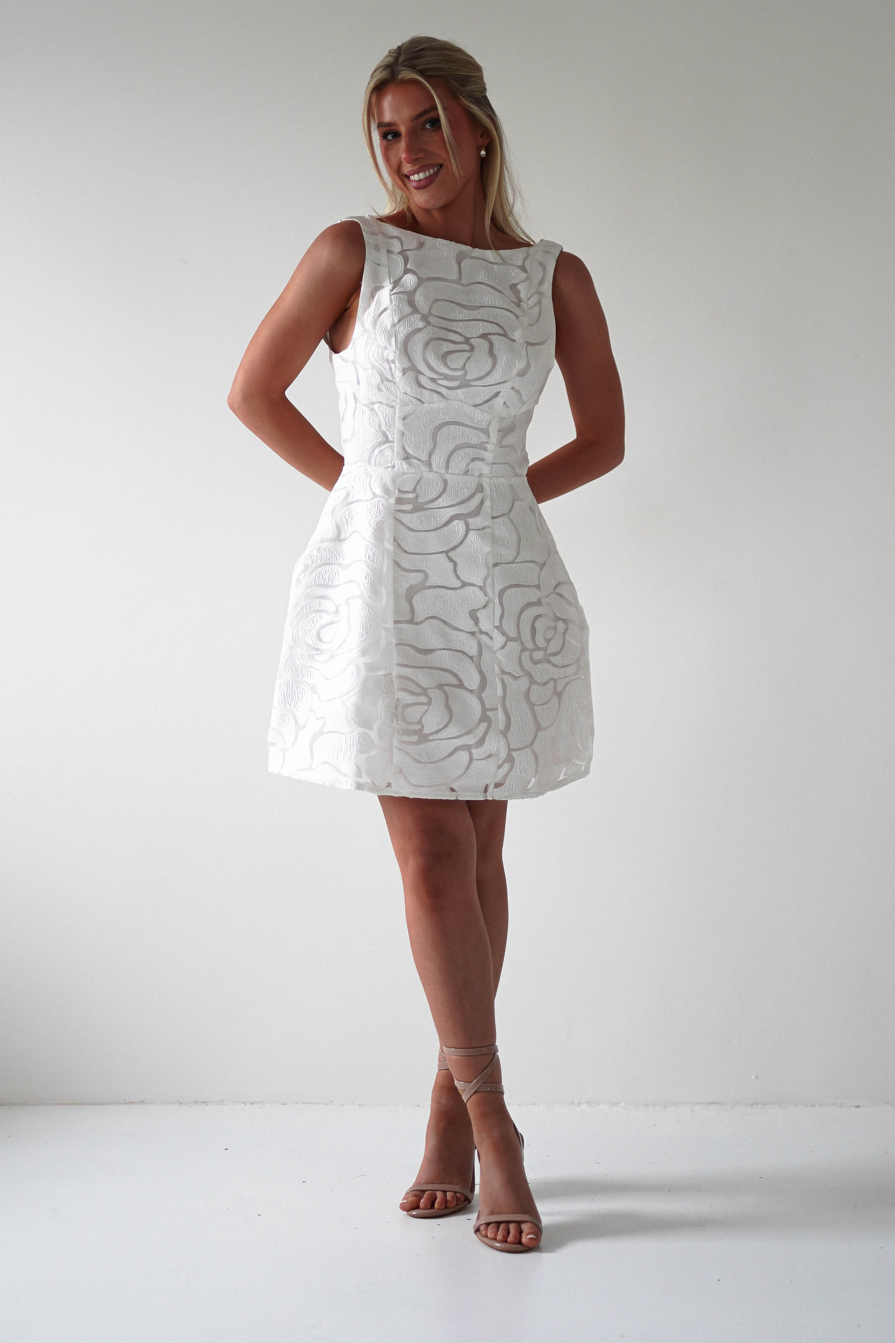 A woman in an embroidered mesh dress poses against a plain white background. Her hands are clasped behind her back, and she smiles. As a perfect choice for weddings or graduations, she wears strappy beige heels, and her long hair is tied back.
