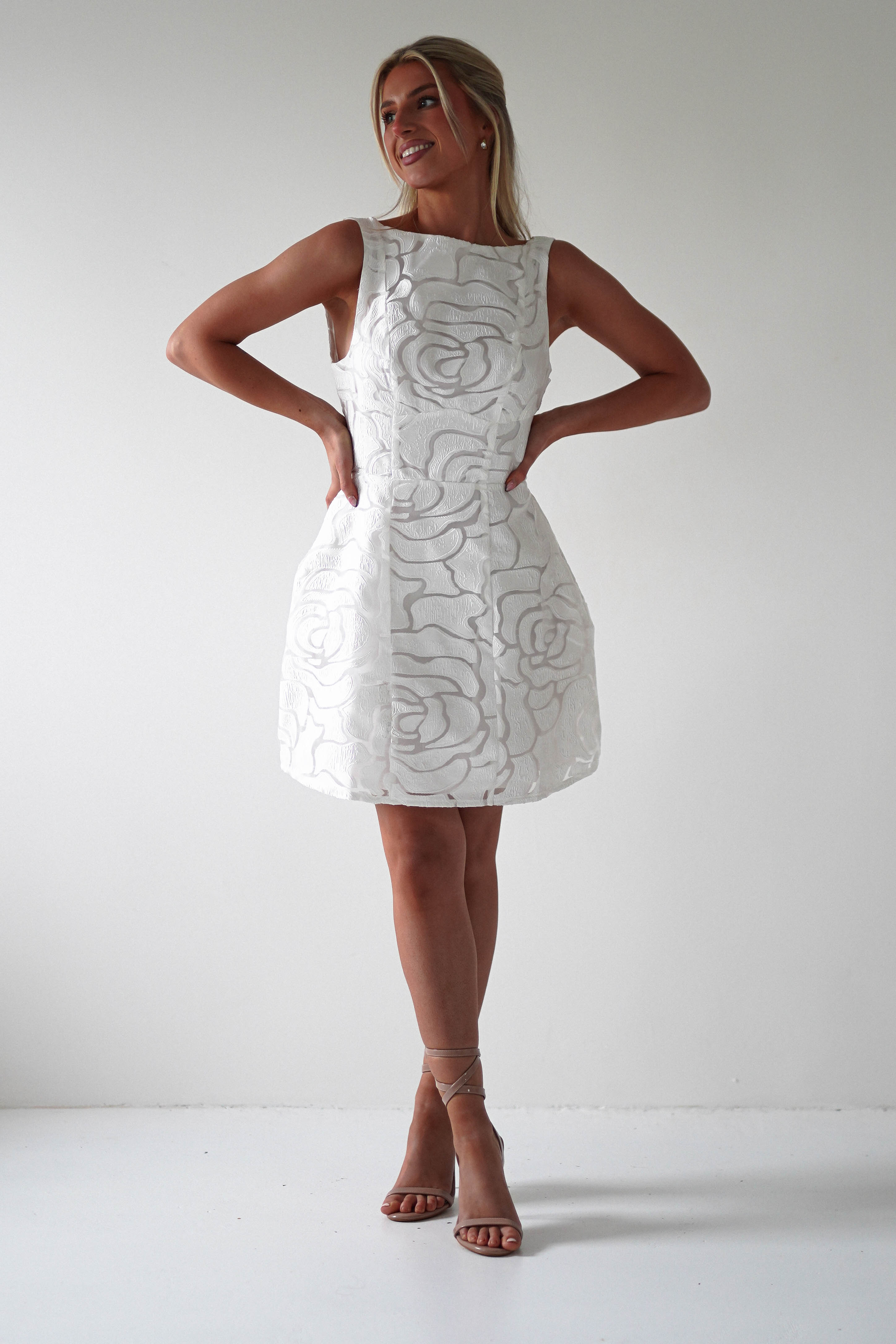 Woman smiling and posing in an embroidered mesh dress with a floral pattern. Perfect for graduations or as a stylish wedding guest, she stands against a plain light-colored background, wearing strappy high-heeled sandals.