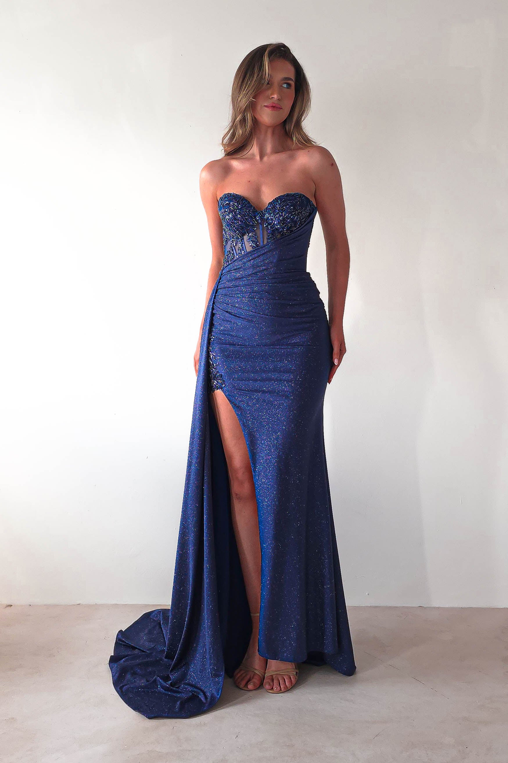 A woman in a glitter gown with a sweetheart neckline and high slit stands against a plain background. Her long hair cascades over the sparkly, strapless blue fabric, complementing her strappy sandals—perfect attire for formal events.