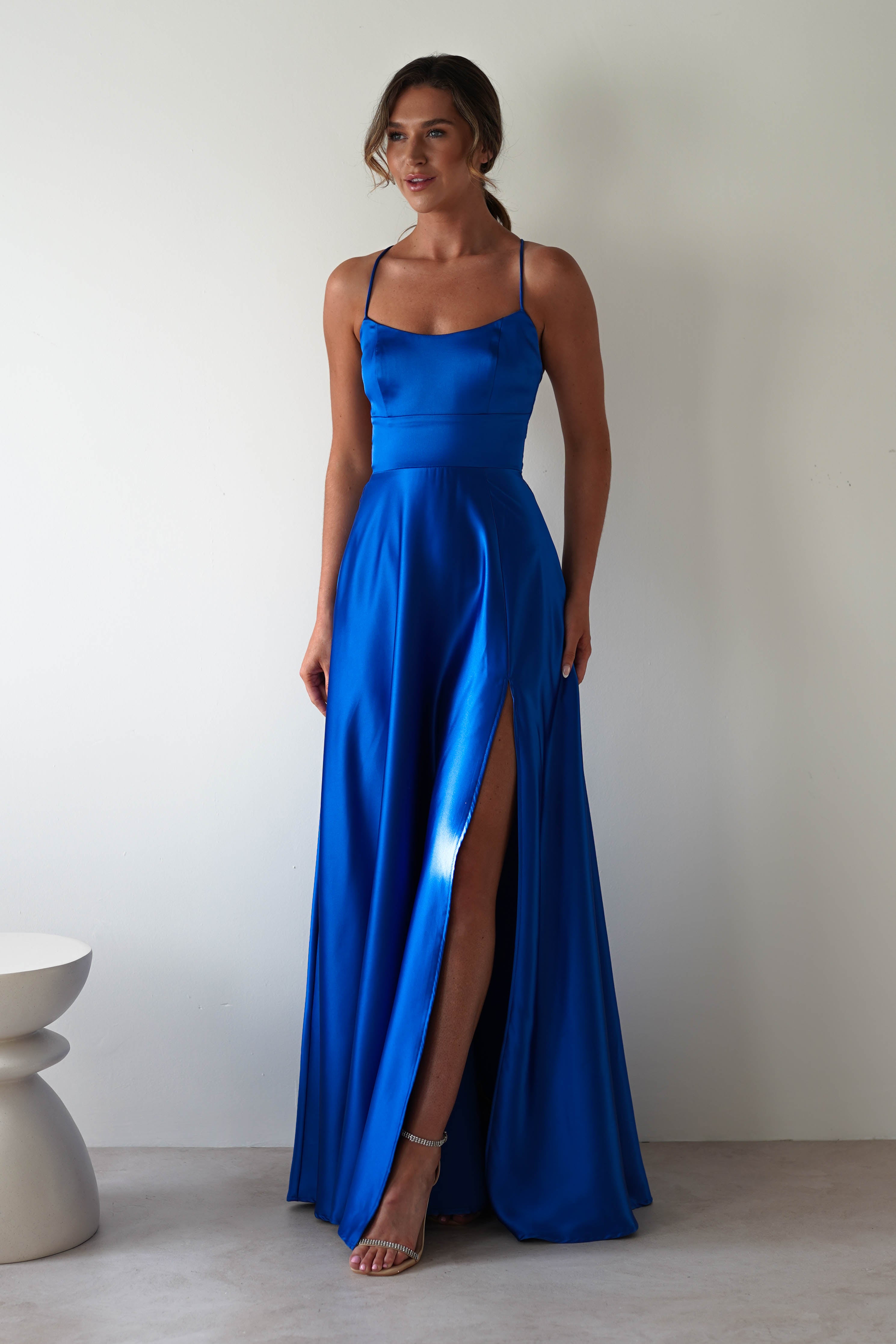 A woman stands against a plain background, elegantly wearing the Leelie Silky Satin Maxi Gown in royal blue. The sleeveless dress features thin straps and a high slit, showing one leg. Her hair is styled up, and she wears strappy high heels. A modern sculptural stool is nearby.