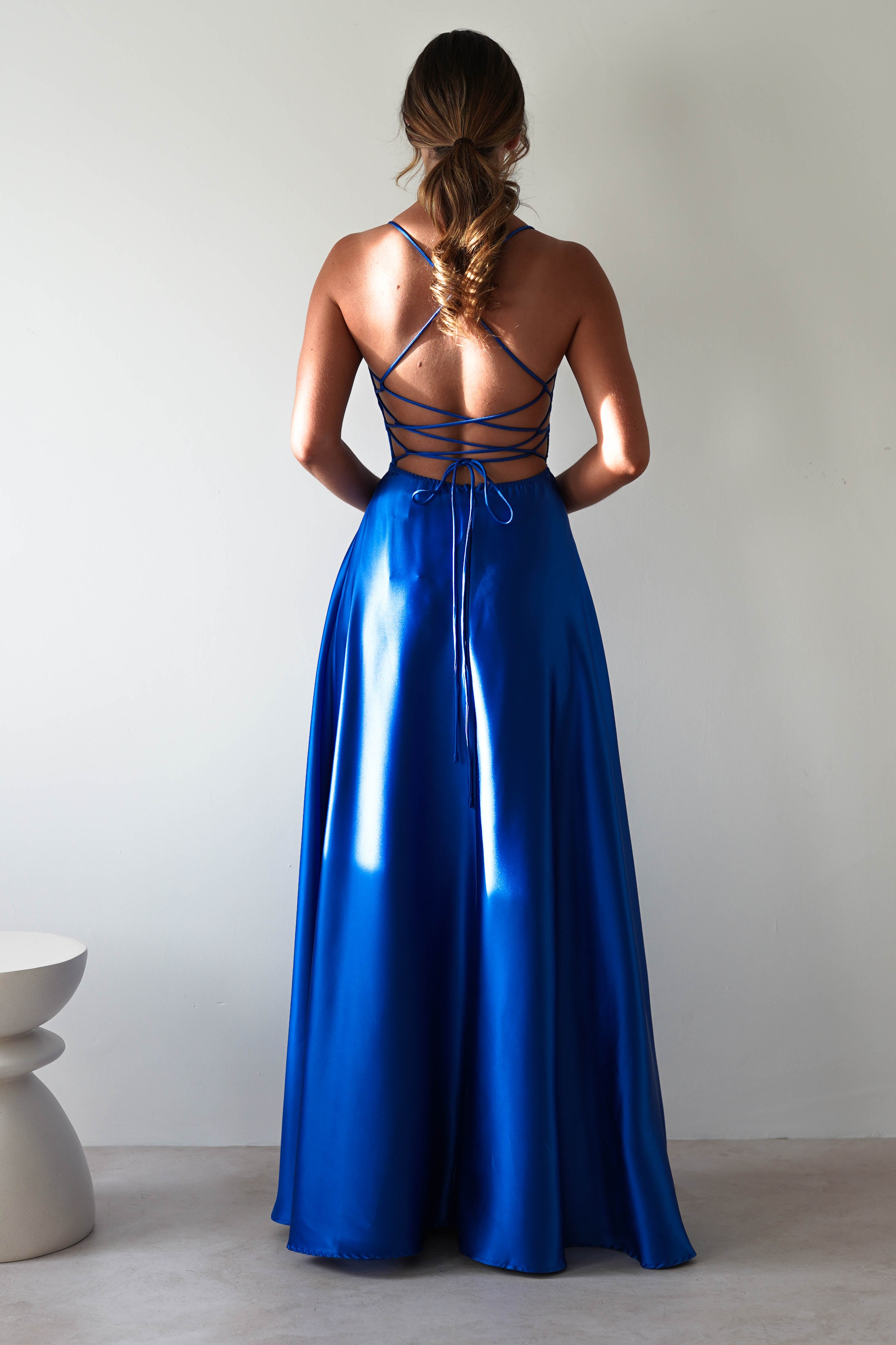 A woman in the Leelie Silky Satin Maxi Gown, royal blue with crisscross straps, faces away. The floor-length dress flows elegantly, and her hair is styled in a loose braid. She stands near a minimalist white stool against a plain gray wall.