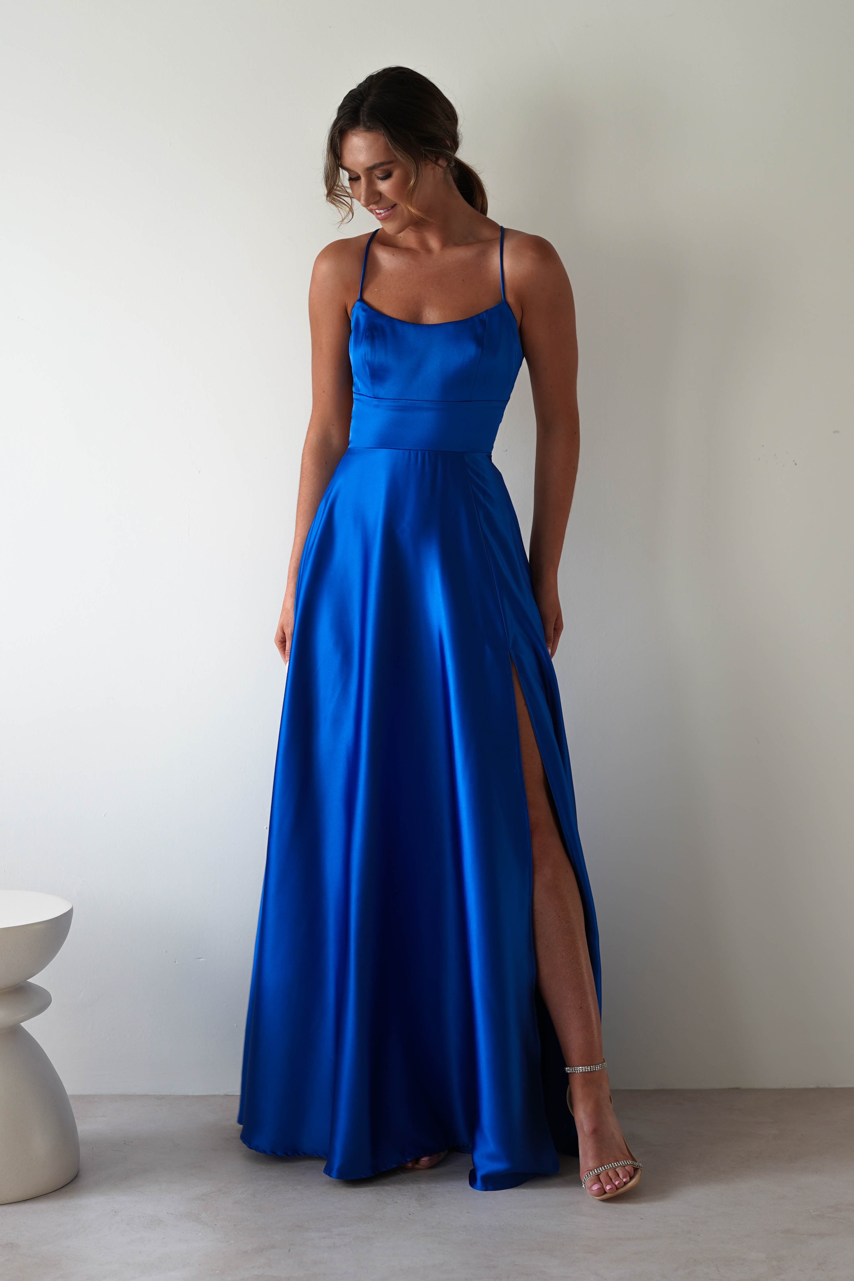 A woman in a Leelie Silky Satin Maxi Gown in royal blue poses against a plain wall. Featuring thin straps and a high slit that reveals her left leg, she stands beside a small, round, white stool with her hair styled back.