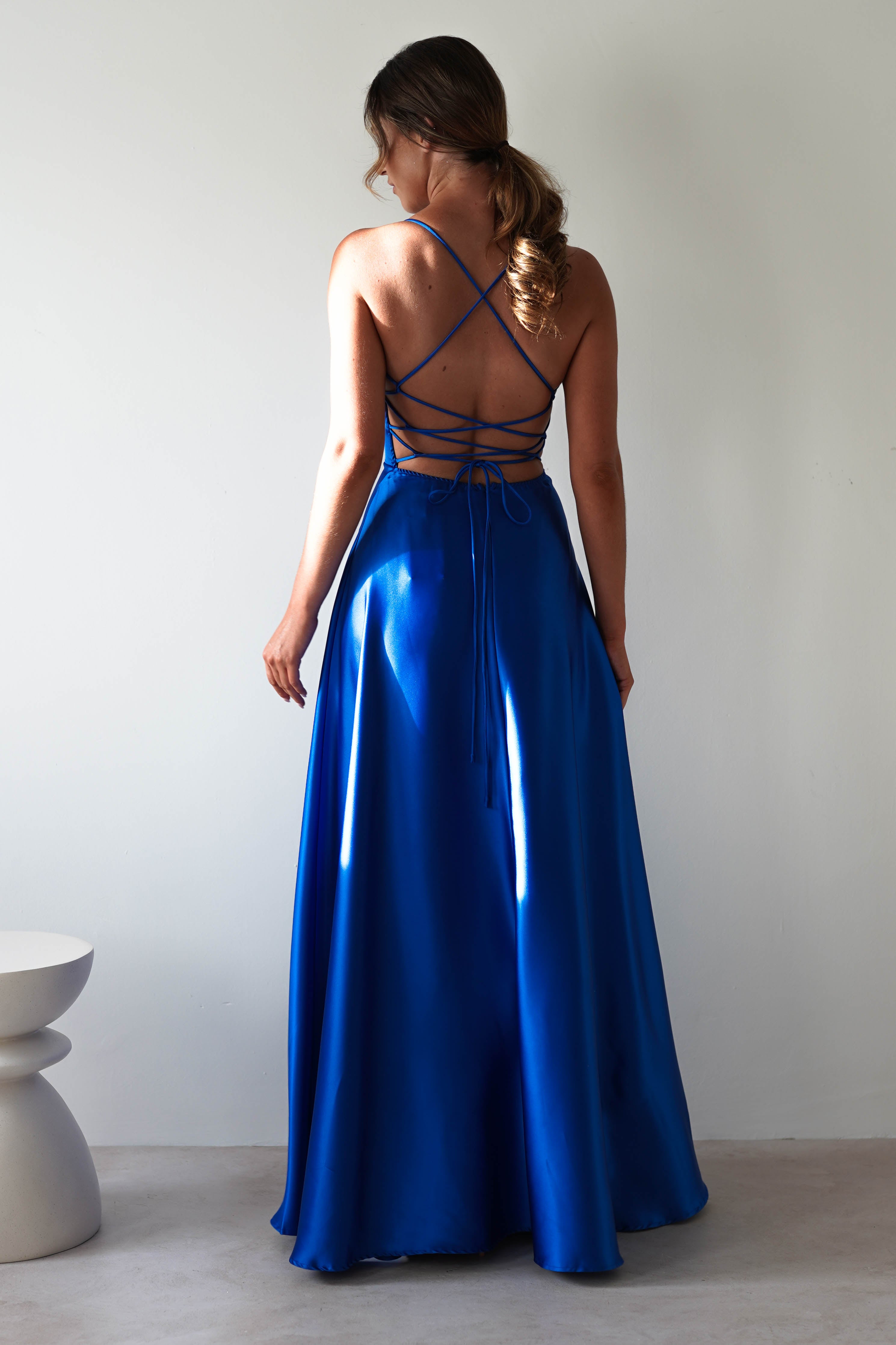A woman wears the Leelie Silky Satin Maxi Gown in royal blue. The gown features a criss-cross strap back design and flowing skirt, embodying classic prom elegance. She stands against a plain white background with a small white stool nearby.