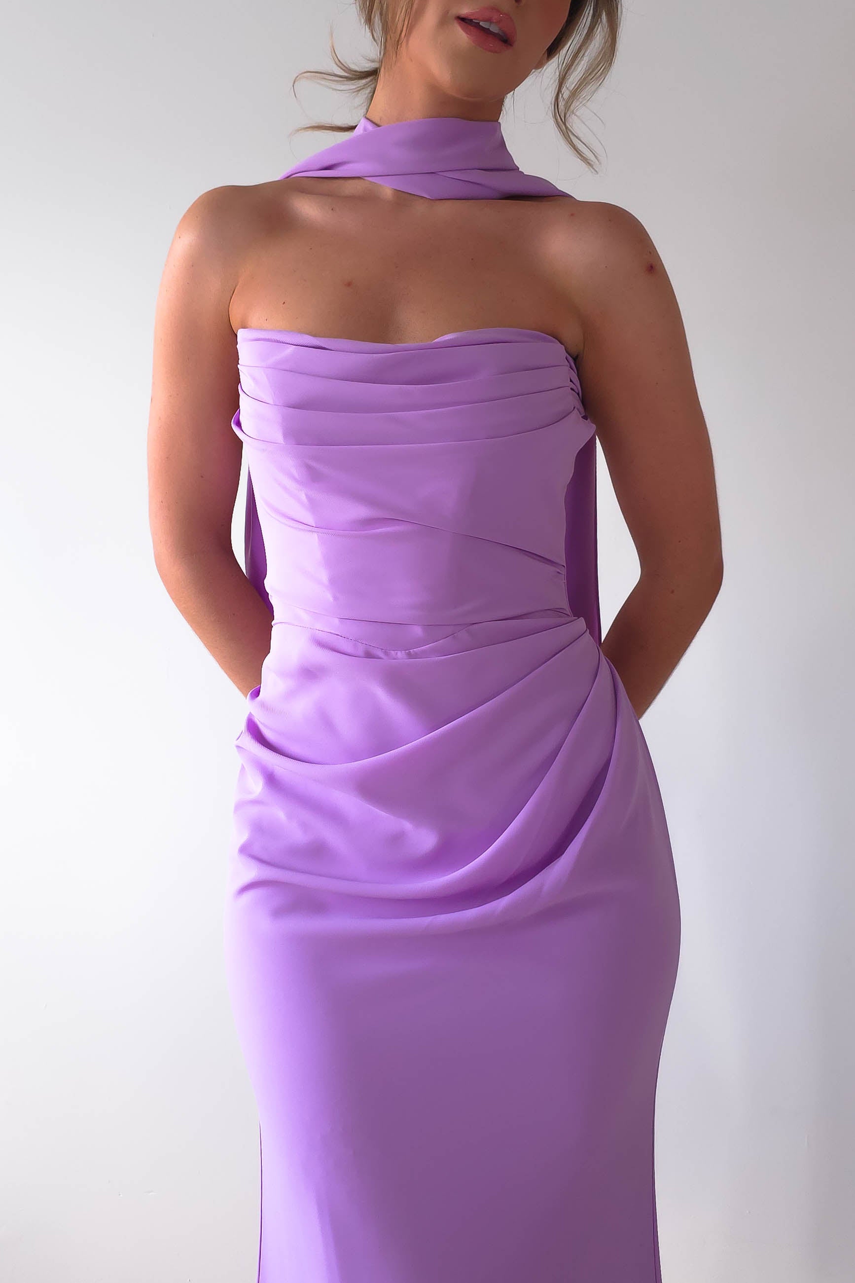 A woman wearing the Lisa Multiway Chiffon Gown, a strapless design in lilac with a draped finish and matching neck scarf. The backdrop is a simple white, highlighting the gowns versatile charm.