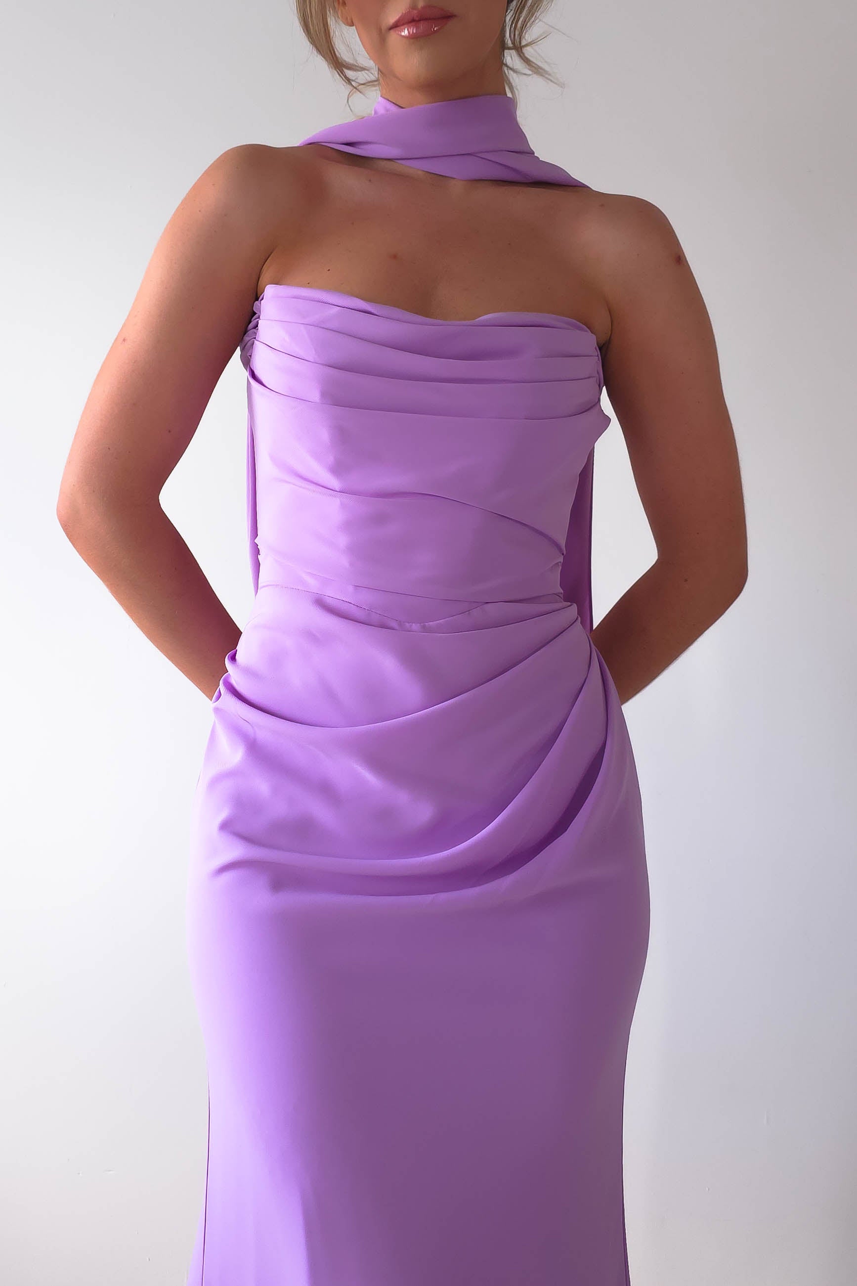A person in a strapless Lisa Multiway Chiffon Gown with elegant draping and versatile ties stands against a plain white background. The lavender gown showcases a fitted silhouette, complemented by a matching neck scarf. The persons face remains unseen.