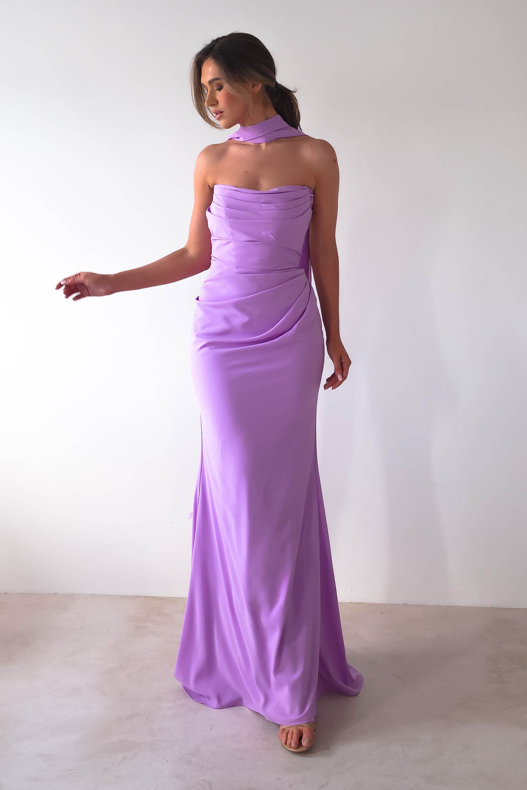 A woman poses in a strapless lavender evening gown, the Lisa Multiway Chiffon Gown, with a matching neck scarf against a plain white background. The dress features elegant draping, complemented by her open-toed shoes.