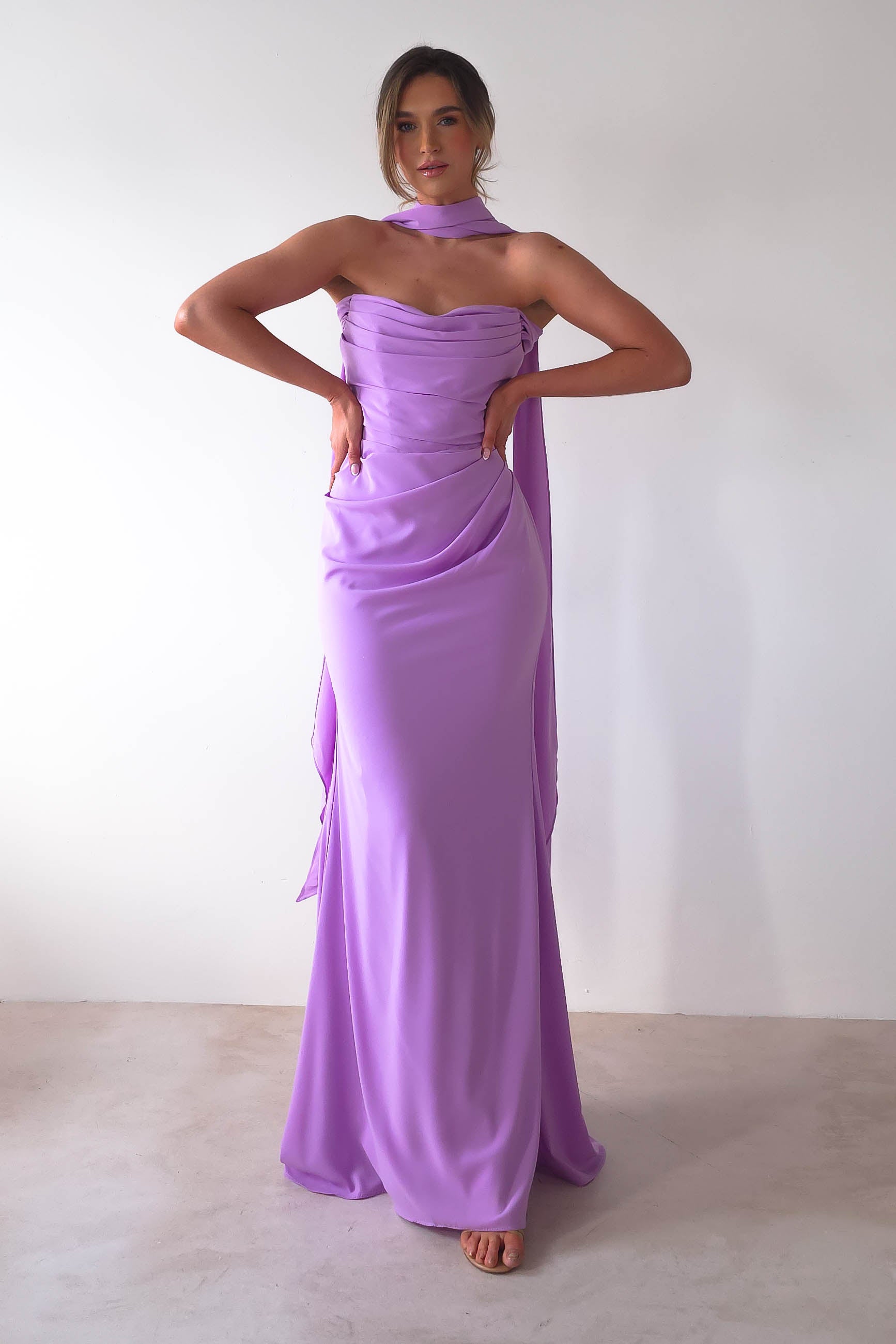 A person poses before a plain backdrop in a strapless Lisa Multiway Chiffon Gown, draped with an elegant scarf. Their hands rest confidently on their hips as they gaze towards the camera.