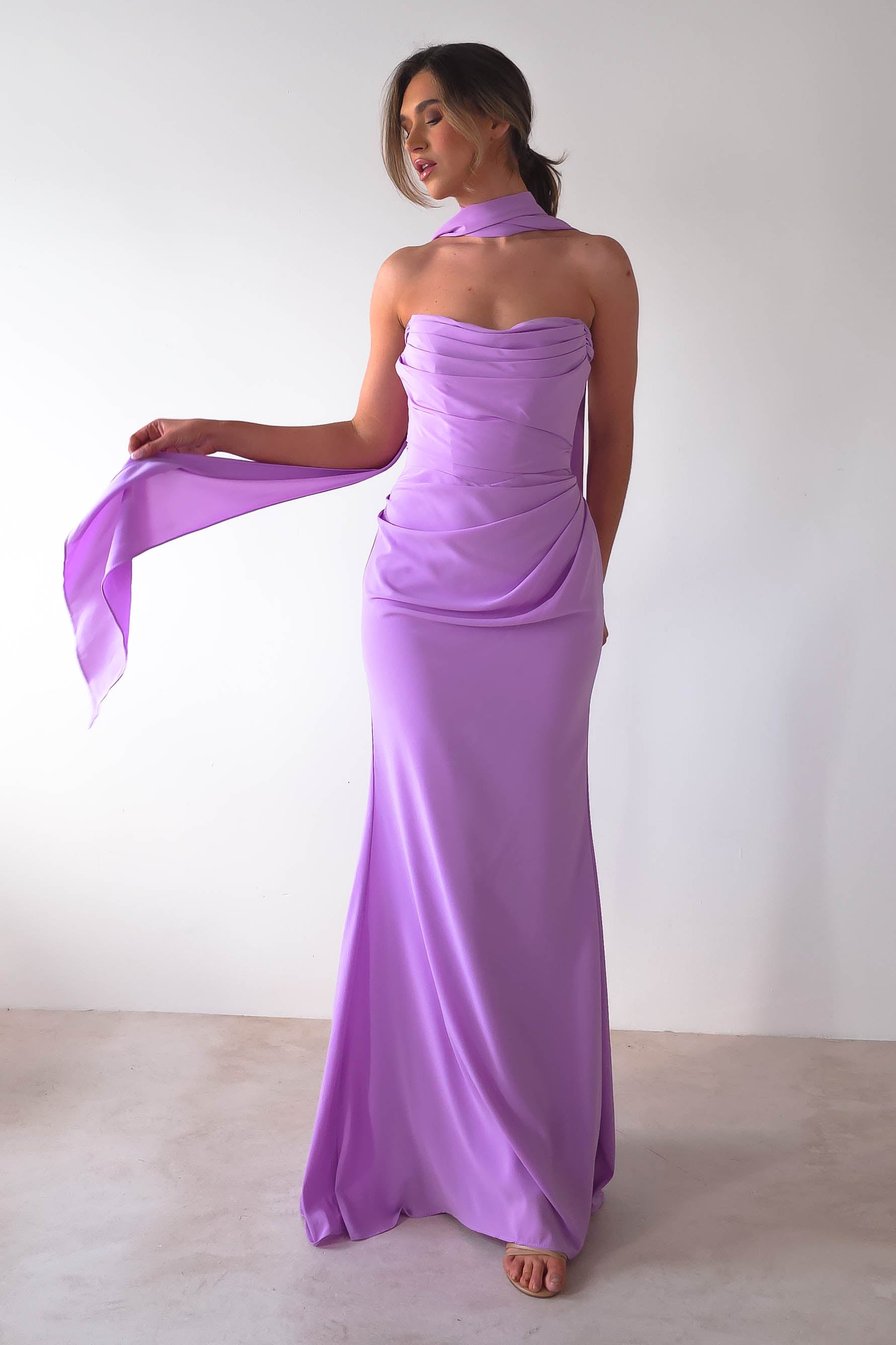 A woman stands against a plain white background wearing the Lisa Multiway Chiffon Gown in lavender. Its strapless design is fitted and drapes elegantly to the floor with versatile ties. Holding one end of her flowing scarf, she looks poised and serene.