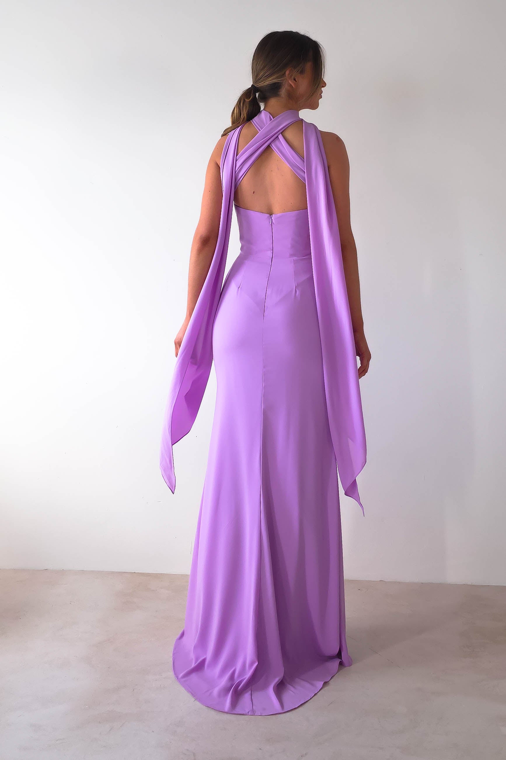 A woman in a floor-length Lisa Multiway Chiffon Gown with crossed straps on the open back and a long drape. The dress flows elegantly towards the floor, showcasing its versatile ties. She stands against a plain white wall, facing away.