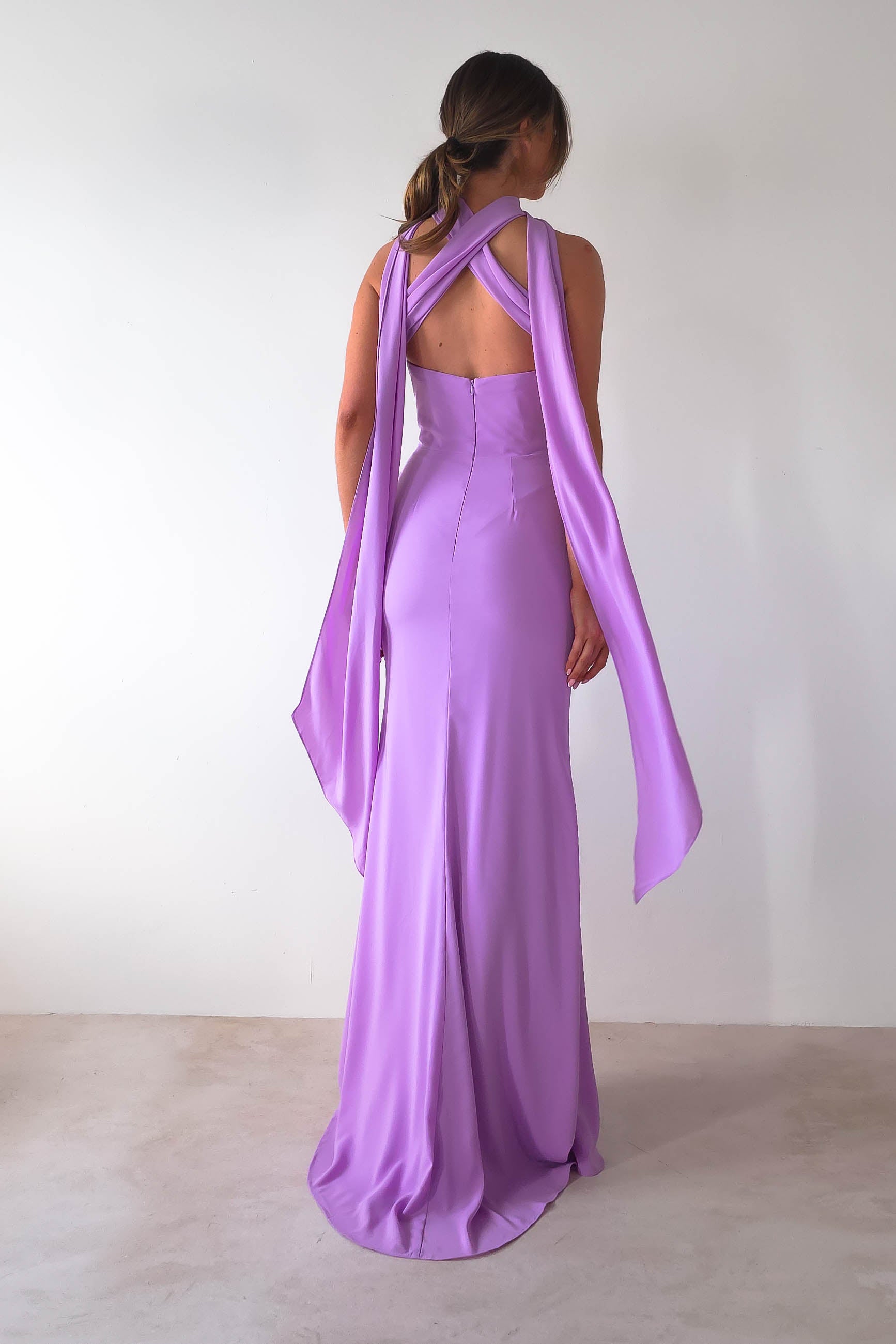 Woman in a lavender Lisa Multiway Chiffon Gown with long, flowing fabric details on the shoulders and back, stands against a white background. The dress features a fitted silhouette and versatile ties for a personalized style. Her hair is tied back, showcasing the gowns elegance.
