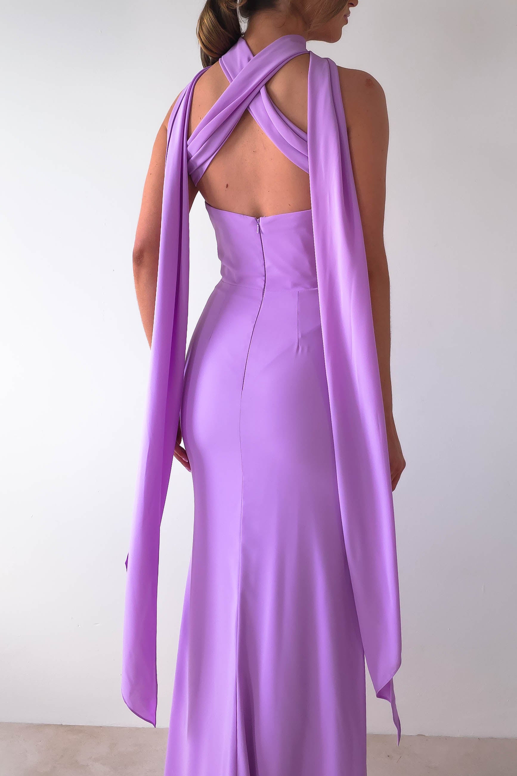 A woman in a floor-length lavender gown, reminiscent of the Lisa Multiway Chiffon Gown, flaunts its sleeveless elegance with crisscrossed halter straps and versatile ties that cascade beautifully. She stands gracefully against a plain white background.