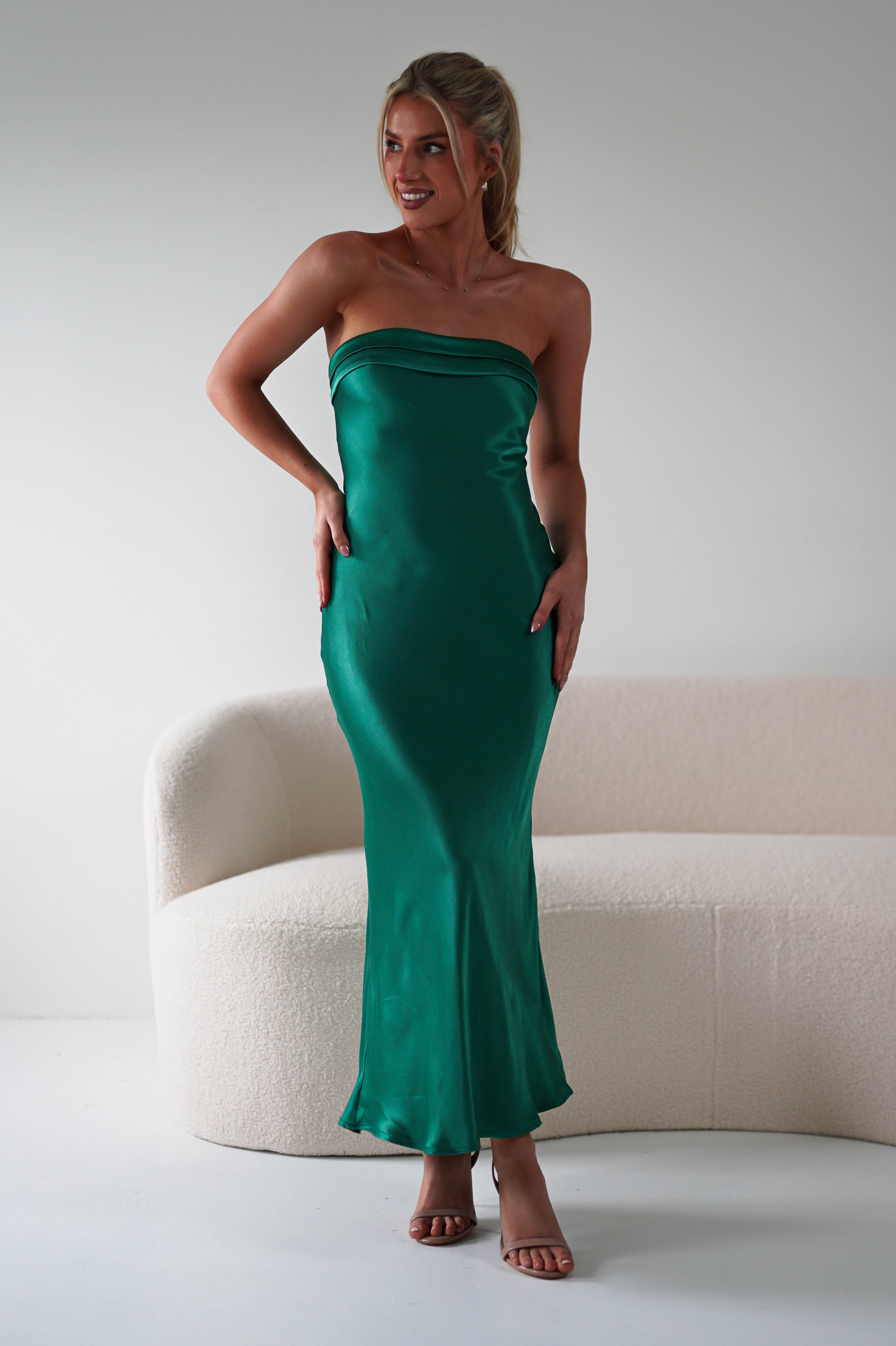 A woman in a strapless midi dress made from super soft fabric stands smiling in front of a white curved sofa. Her hair is tied back, and she wears high-heeled sandals, perfect for wedding guests. The background is minimalist and bright.