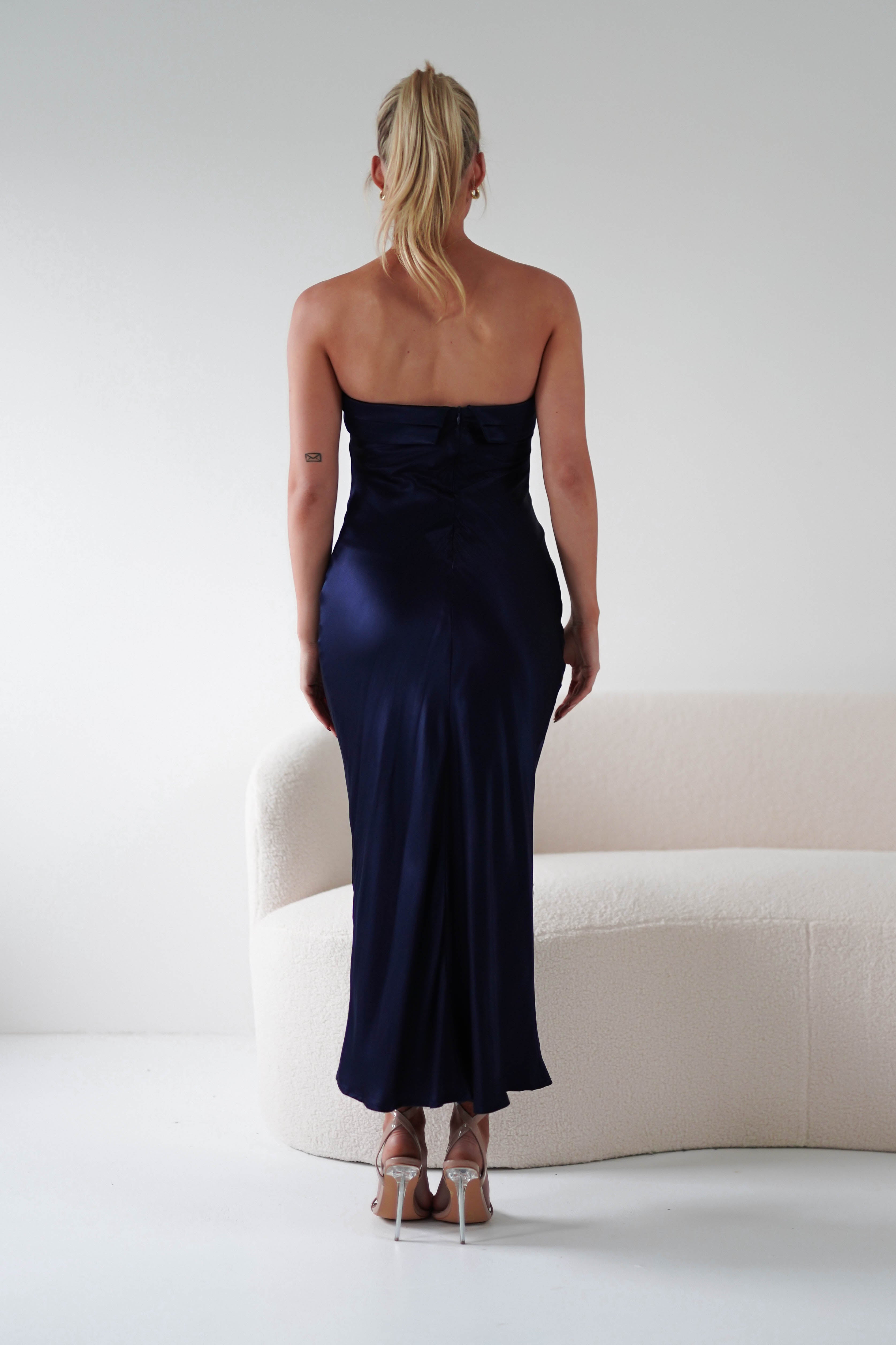 A person with long blonde hair stands facing away, elegantly poised in a strapless navy satin gown made from super soft fabric. They are in front of a white, curved sofa in a minimalistic setting. The fitted gown gracefully flows to the floor, embodying sophistication among wedding guests.