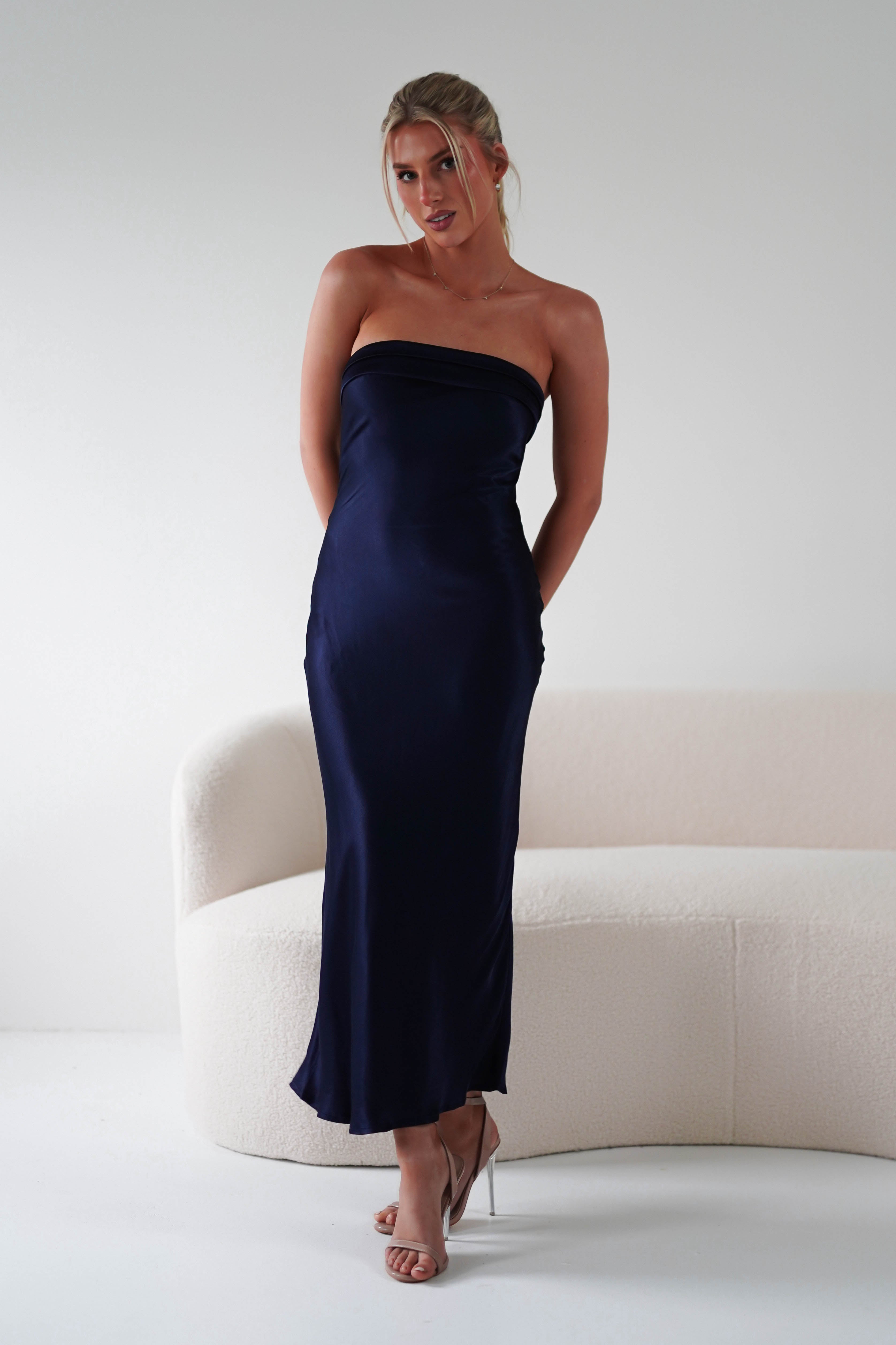 A woman in a strapless dark blue satin midi dress stands gracefully in front of a light-colored curved sofa. Her hair is styled up, and she wears high heels, fitting perfectly among elegant wedding guests. The setting is minimalist with a crisp white background.