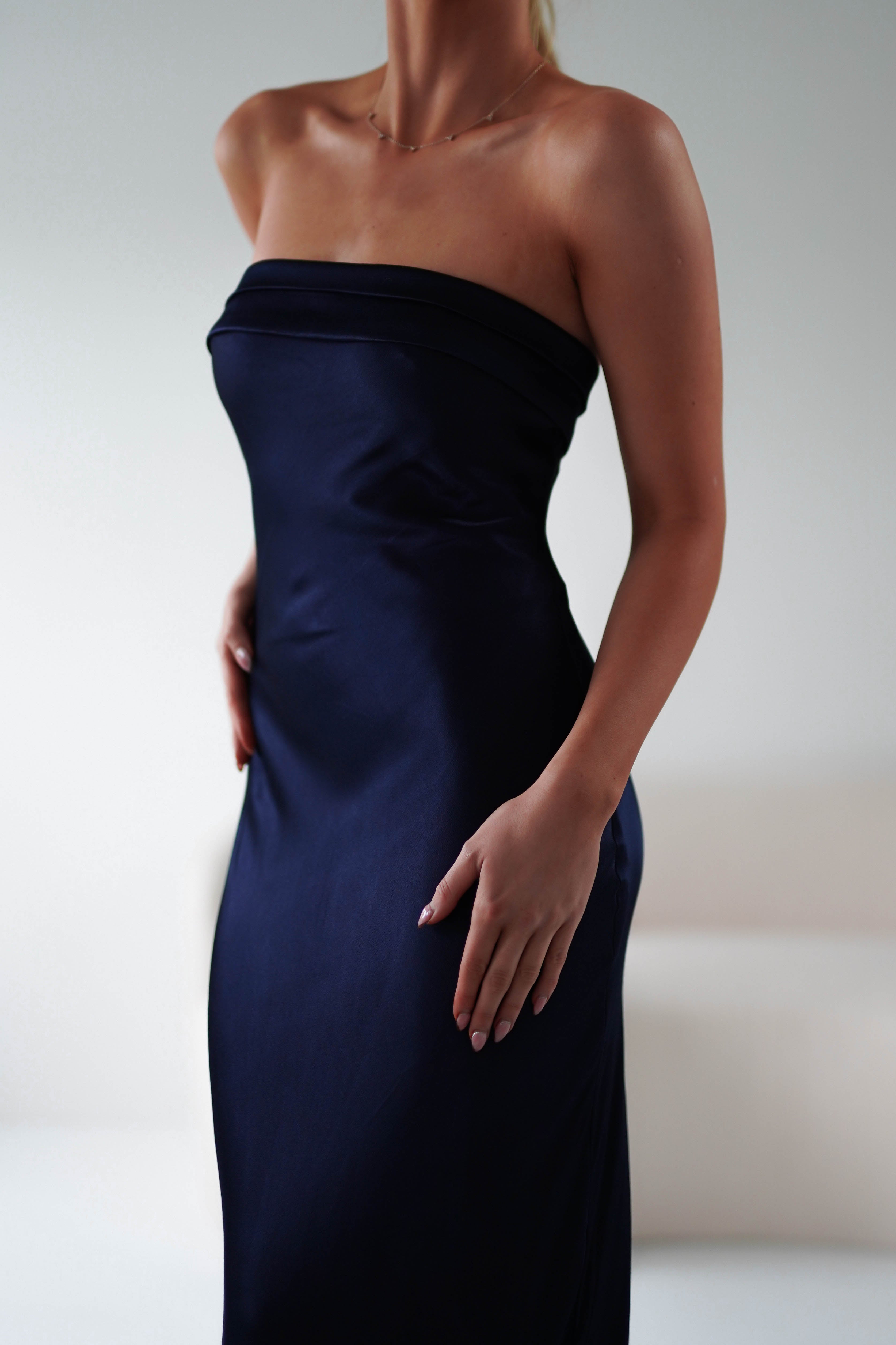 A person wearing a sleek, strapless midi dress in dark blue stands with hands relaxed by their sides. The super soft fabric gently reflects light, highlighting its smooth texture. The background is blurred, drawing focus to the elegant attire perfect for wedding guests.