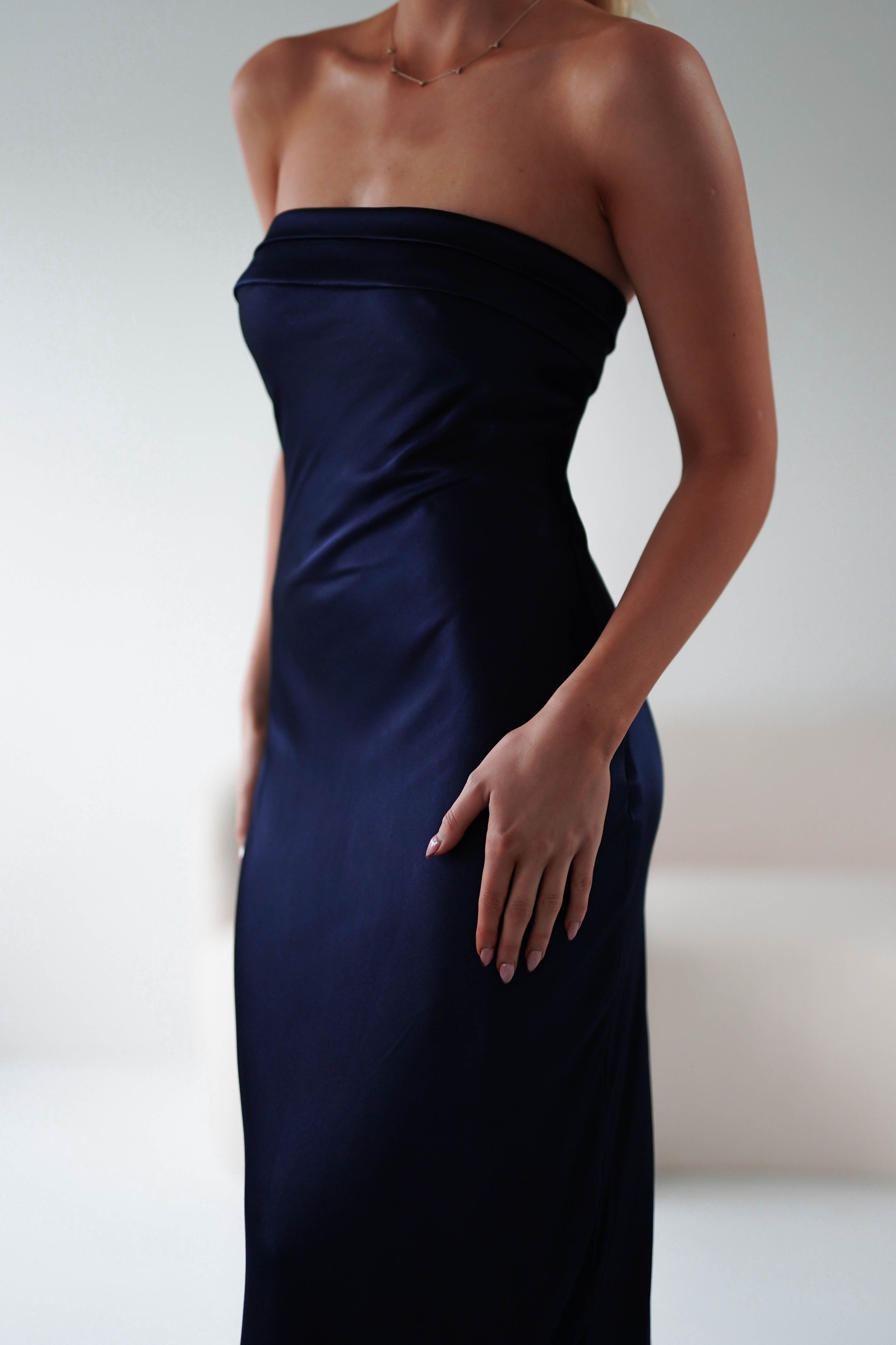 A person wearing an elegant, strapless midi dress in navy blue stands in a softly lit room. The focus is on their torso and the super soft fabrics smooth texture, while the background is softly blurred, creating a perfect setting for wedding guests.