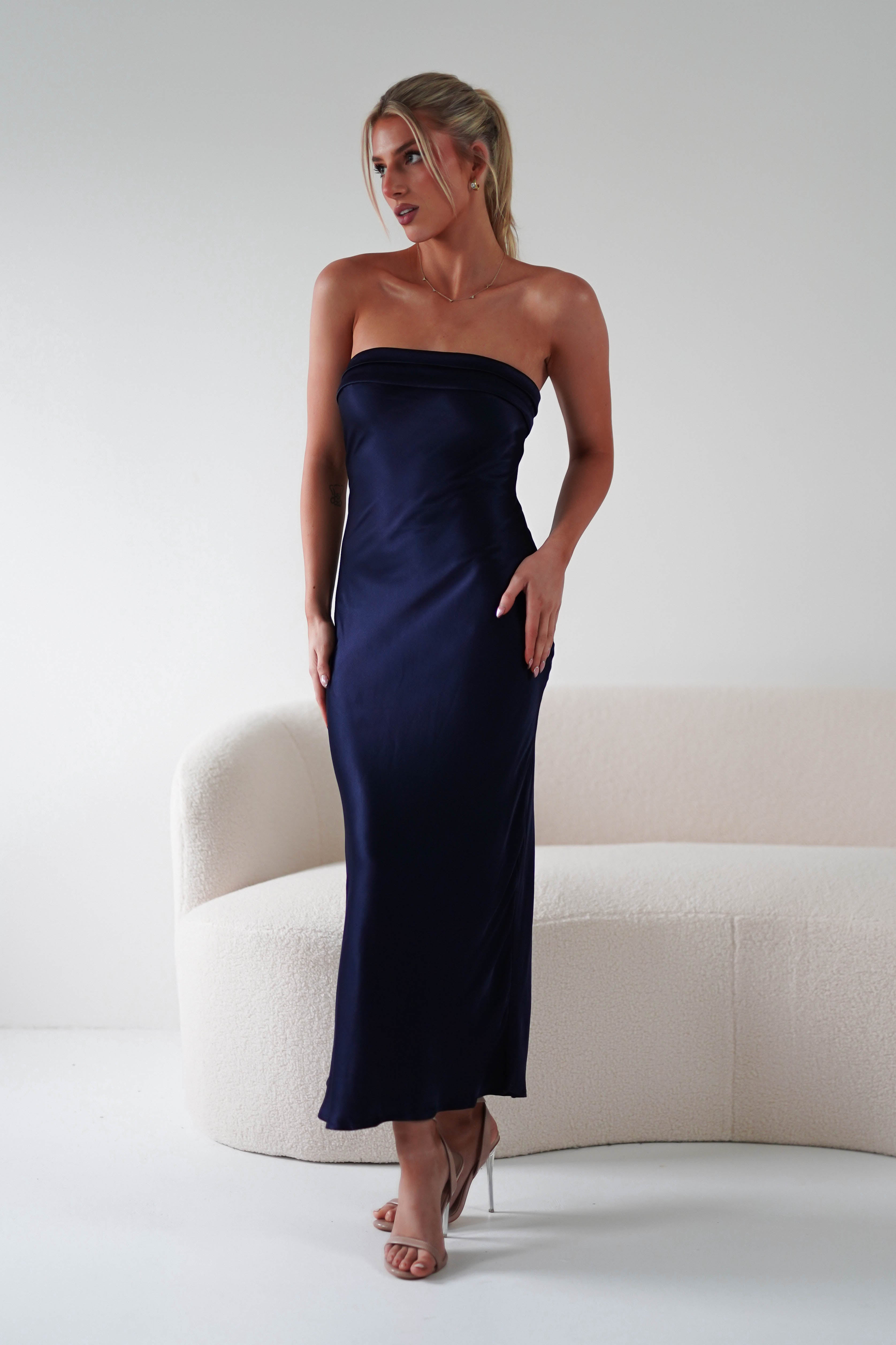 A woman in a navy blue strapless midi dress, made of super soft fabric, stands near a white curved sofa. Her blonde hair is elegantly tied back and shes wearing nude heels. The setting is minimalistic, bathed in soft, diffused light—perfect for wedding guests to admire.