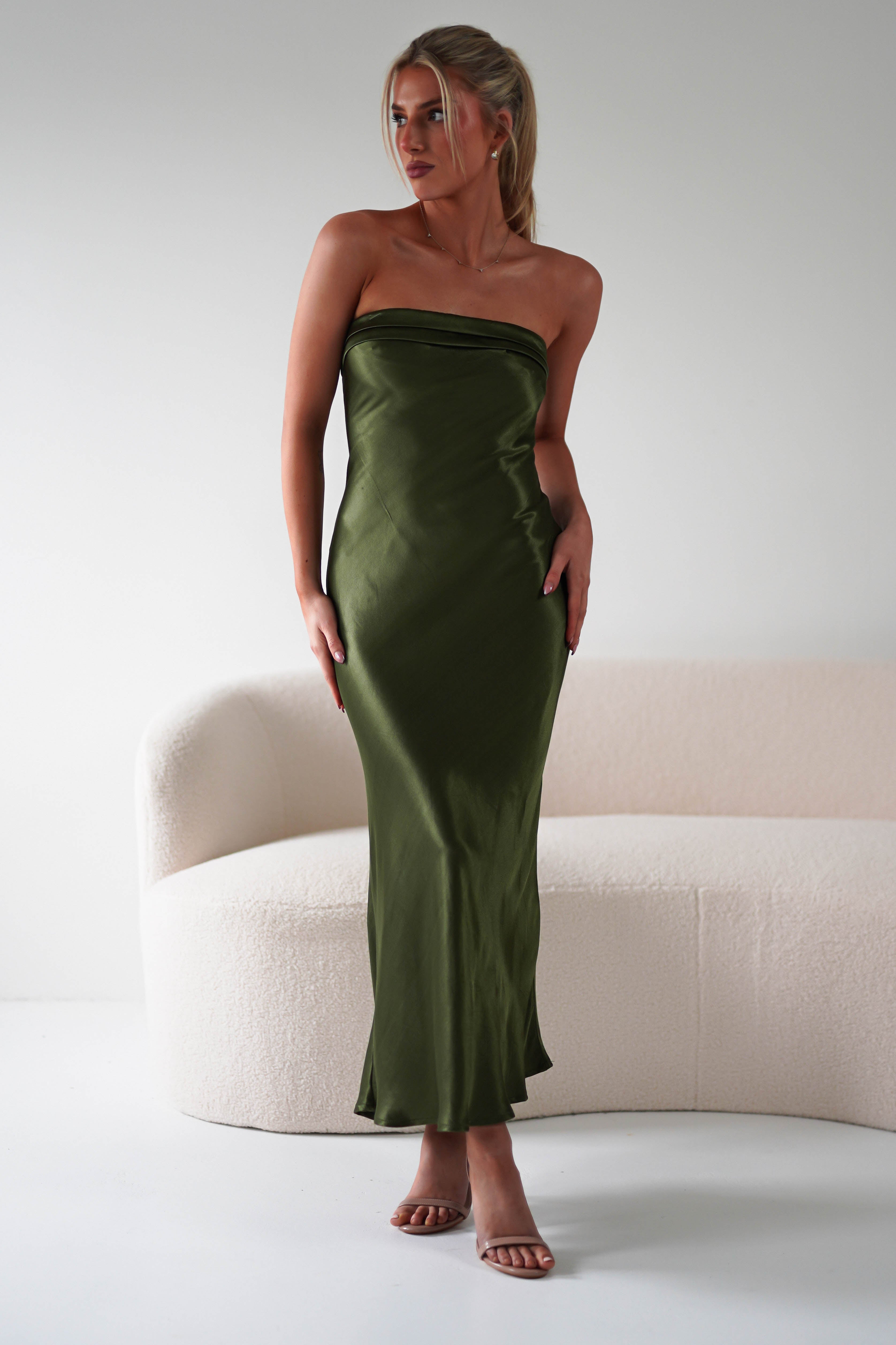 A woman wearing a strapless midi dress in olive green super soft fabric stands in front of a light-colored curved sofa in a minimalistic room. She has blonde hair styled in a ponytail and is wearing tan open-toe heels—perfect for wedding guests.