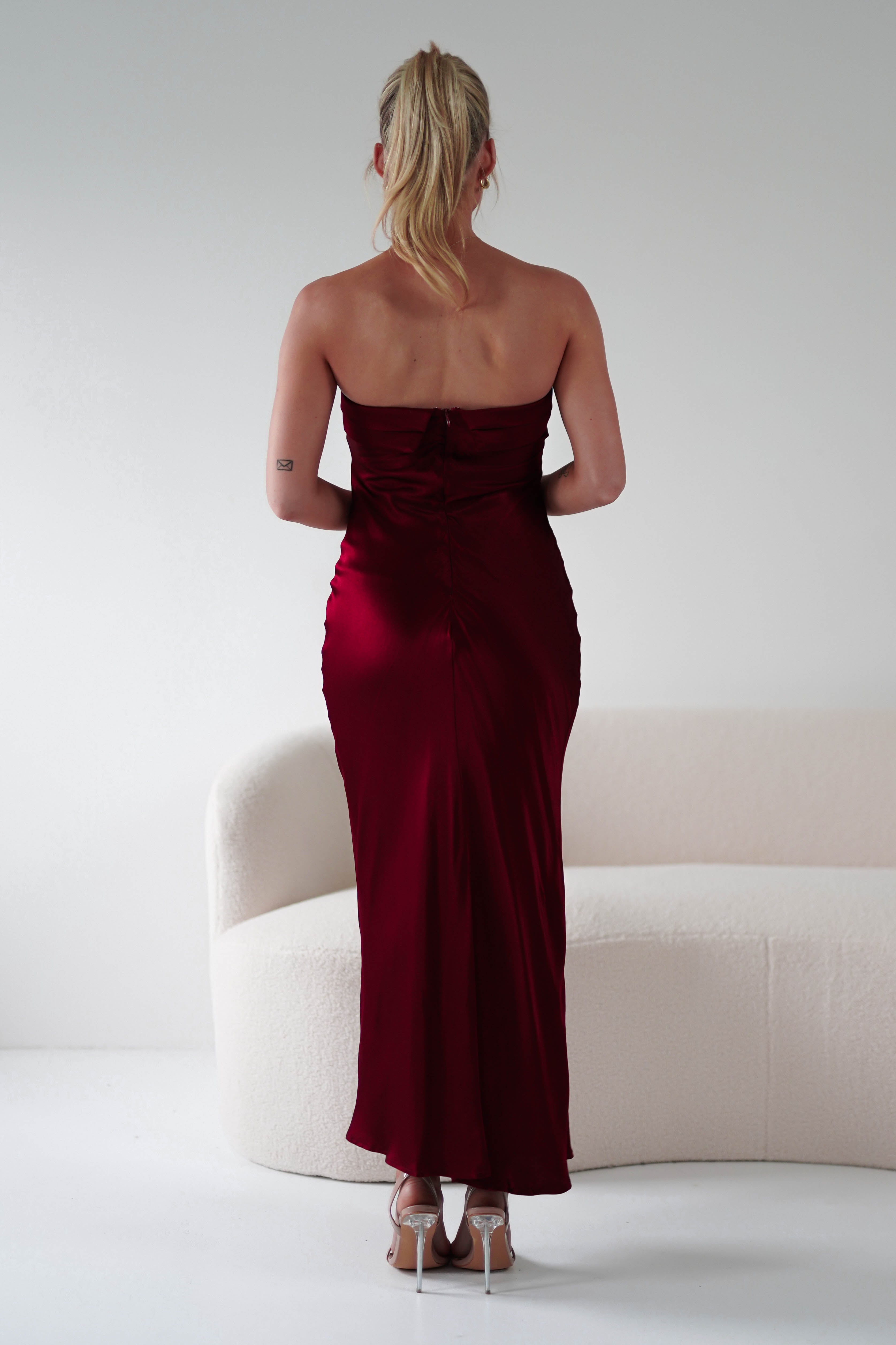 Rear view of a person with long blond hair in a strapless midi dress, exquisitely tailored with super soft fabric, standing in front of an off-white, curved sofa. They are wearing high heels and have a tattoo on their upper right arm. The setting is minimalistic and well-lit.
