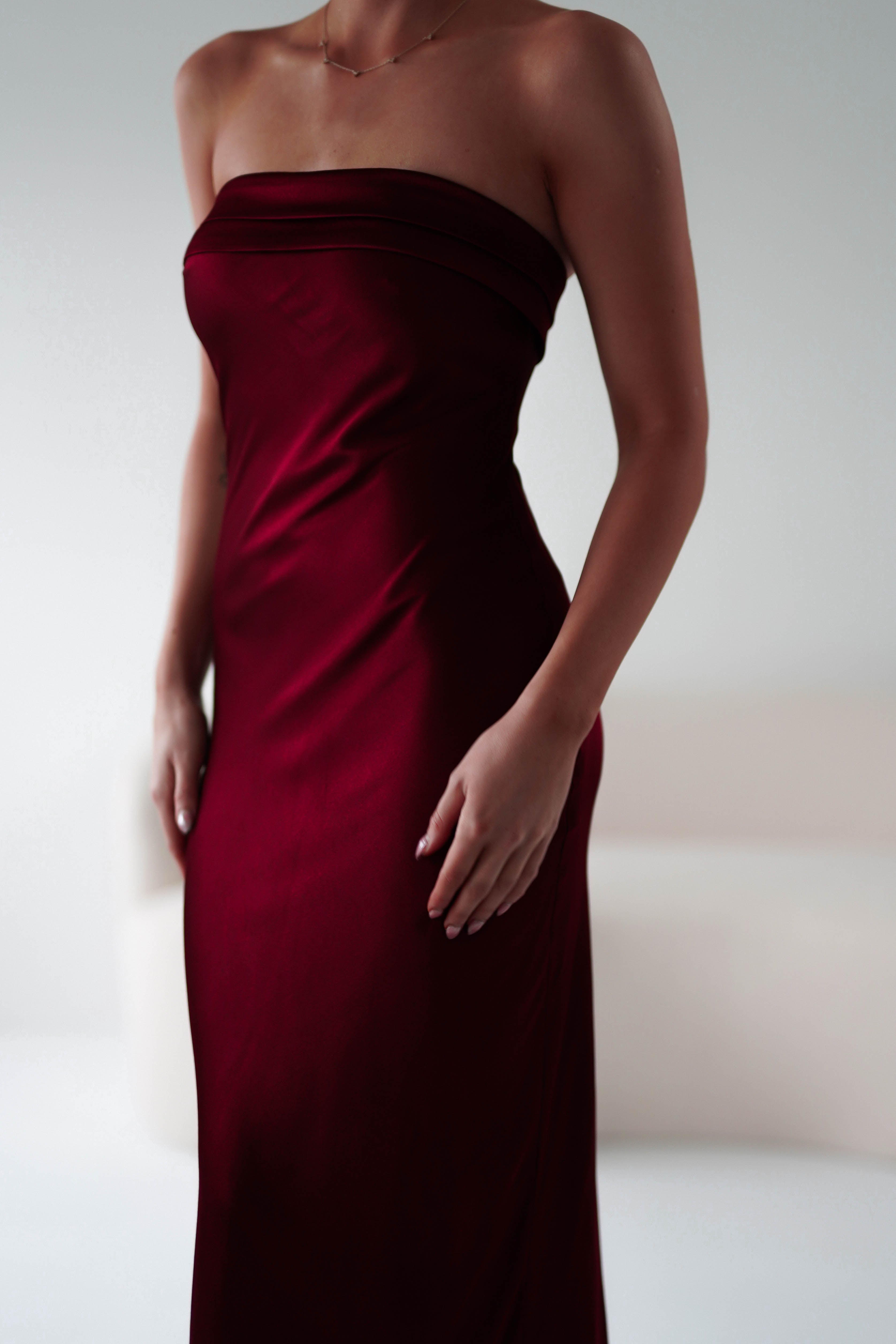 A person wearing a strapless midi dress in form-fitting burgundy stands in a softly lit room. The dress, perfect as a wedding guest dress, boasts super soft fabric and their hands rest gently at their sides amid a blurred, neutral-colored background.