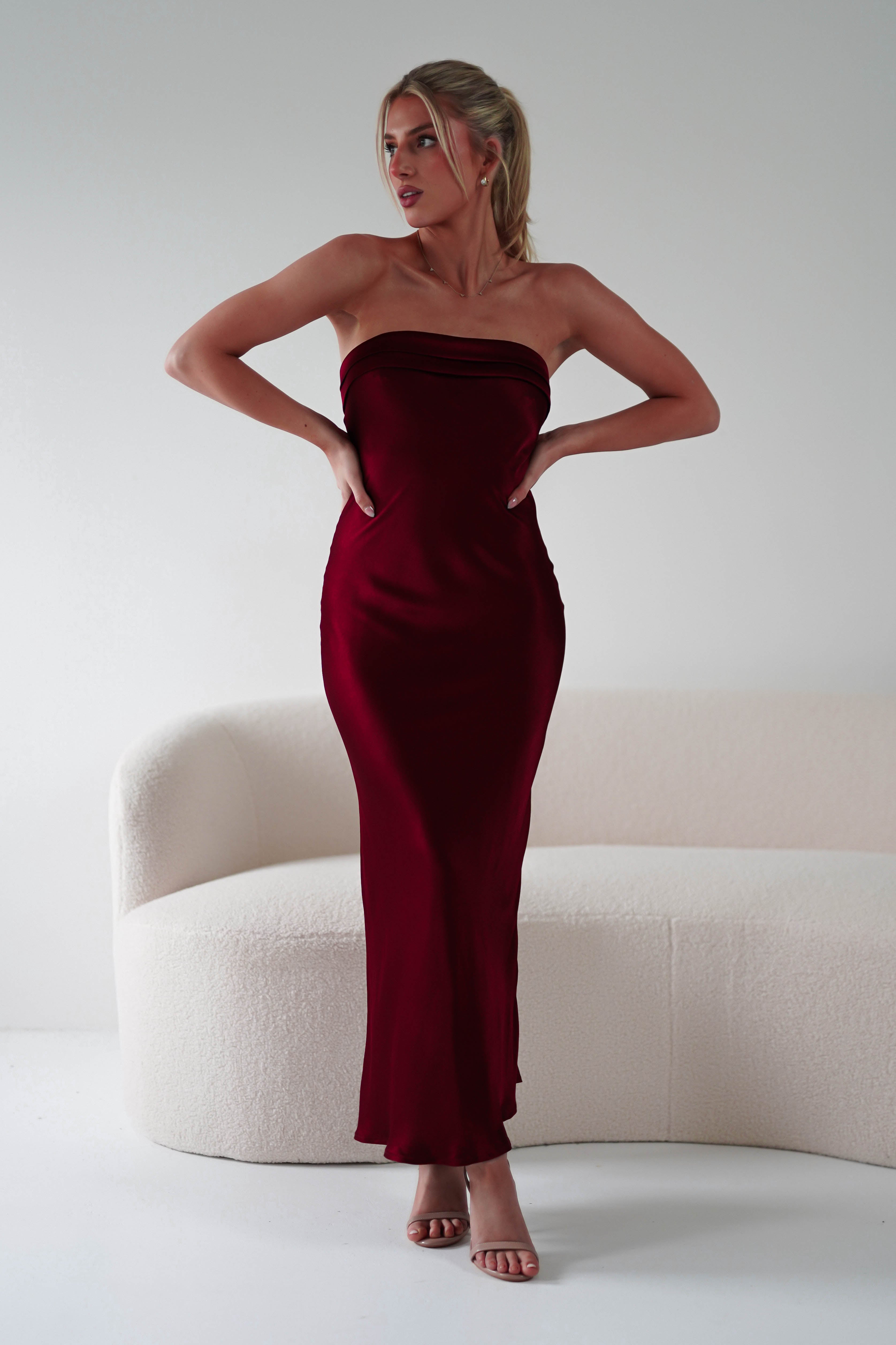 A woman in a strapless, midi burgundy satin gown, perfect as a wedding guest dress, stands with hands on hips in front of a modern, curved white couch. Her hair is styled in a ponytail and she wears high heels. The setting is minimalistic with a plain background.