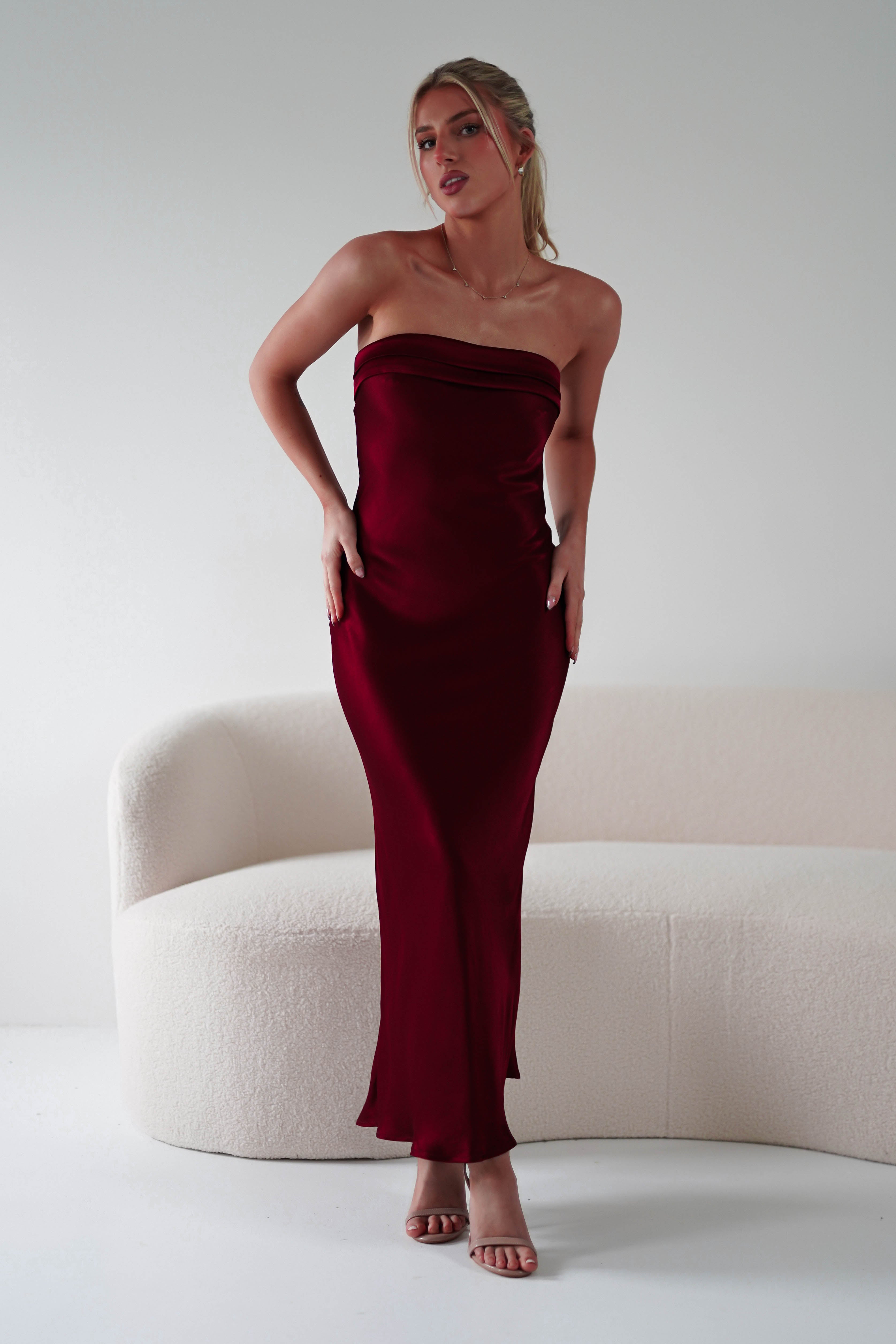 A person in a strapless midi red gown, crafted from super soft fabric, stands in front of a white, modern couch. The sleek and elegant design makes it perfect as a wedding guest dress. They pose with one hand on their hip and the other by their side, wearing high-heeled sandals.