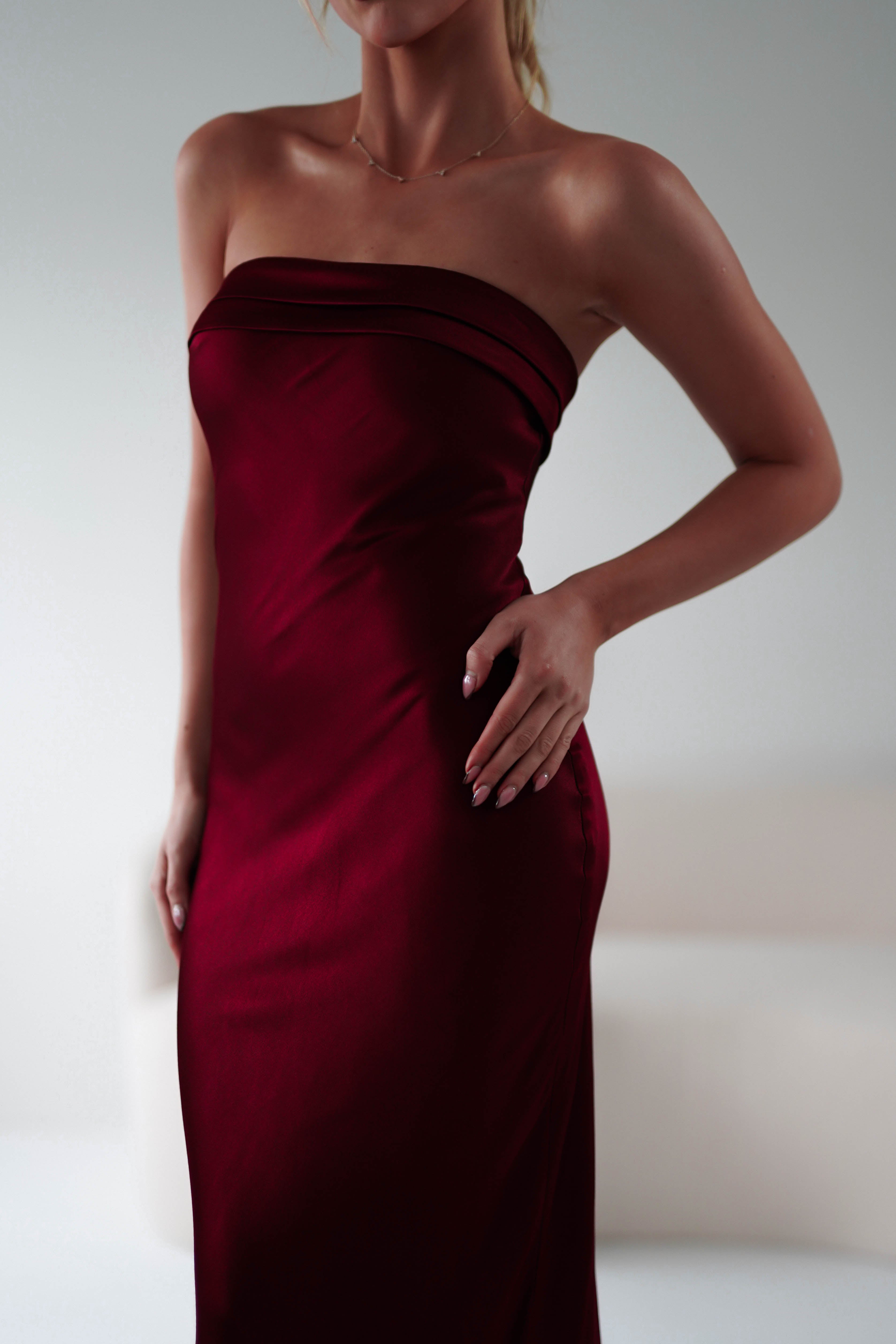 A person in a strapless midi burgundy dress poses with one hand on their hip. The softly lit background highlights the wedding guest dresss rich color and super soft fabric.