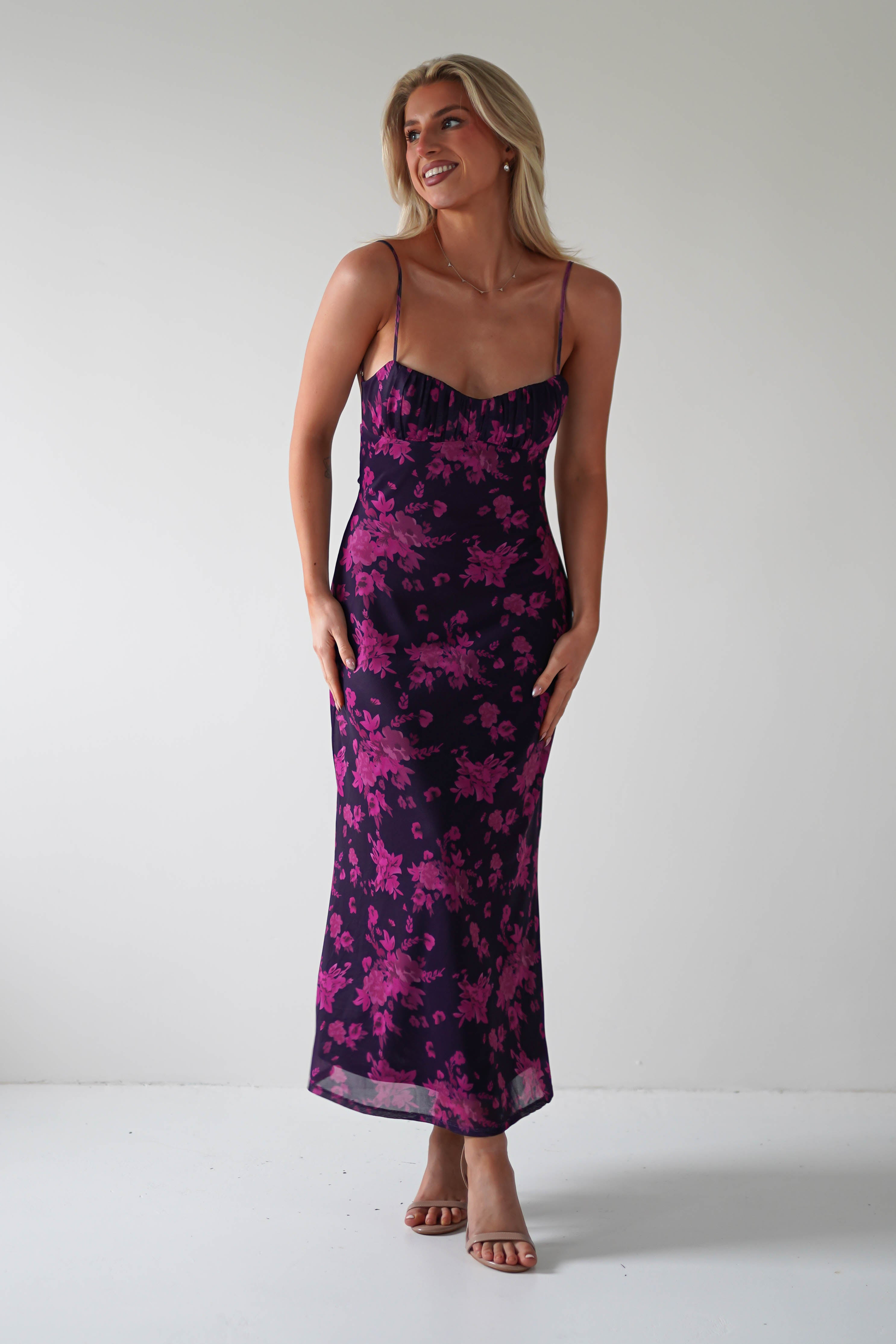 A person is smiling warmly, standing barefoot against a stark white backdrop. Theyre dressed in a sleeveless open back black dress with pink floral patterns flowing gracefully to their ankles. Their long, light hair cascades gently over the elegant ensemble.