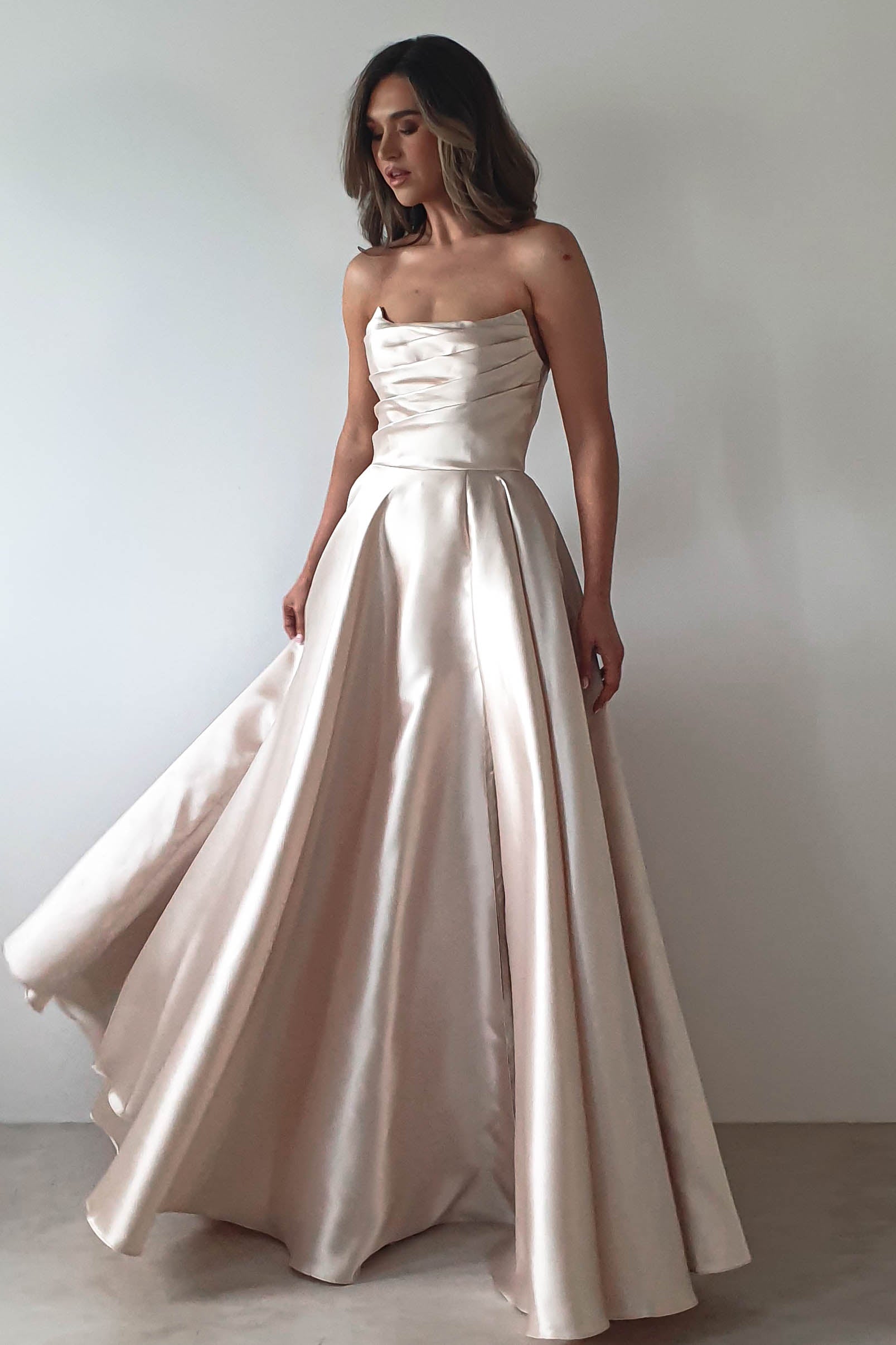 A woman in a strapless, flowing satin gown stands against a plain background. The dress features a fitted bodice with a slight sheen and an A-line skirt perfect for a formal event. She looks to the side, her hair styled in loose waves.