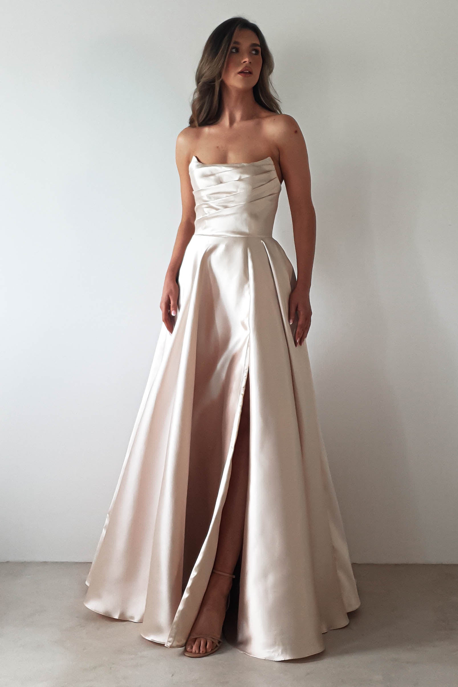 A woman graces a plain white backdrop, clad in a strapless satin gown perfect for a formal event. The floor-length dress showcases an A-line skirt with a subtle slit and features a fitted bodice with delicate pleats. Her long, wavy hair cascades elegantly down her back.