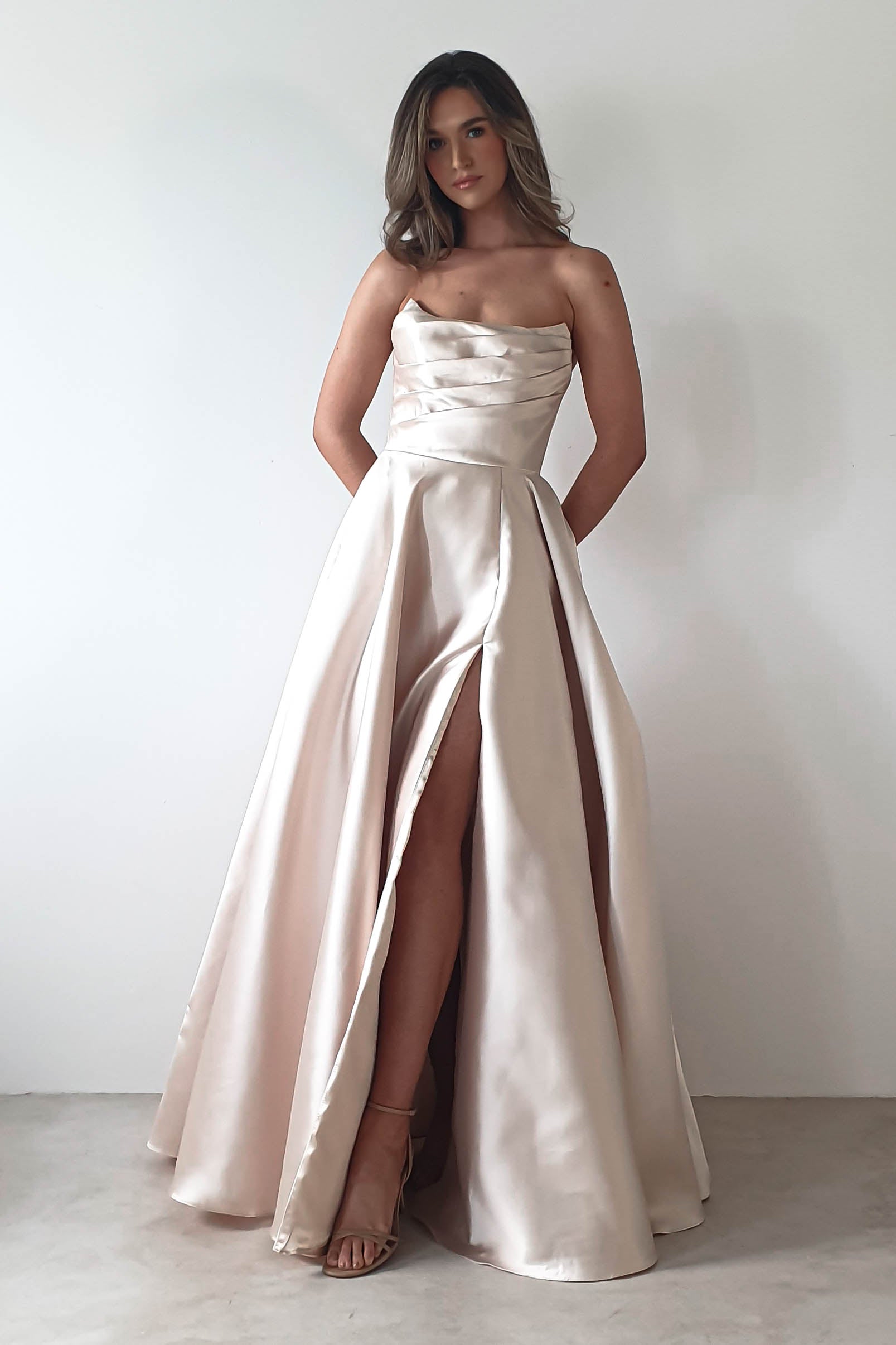 A woman wearing an elegant, strapless satin evening gown stands against a plain white background. Perfect for a formal event, the dress features a fitted bodice and a high slit highlighting her leg. Her hands rest by her sides, and her hair is styled in loose waves.