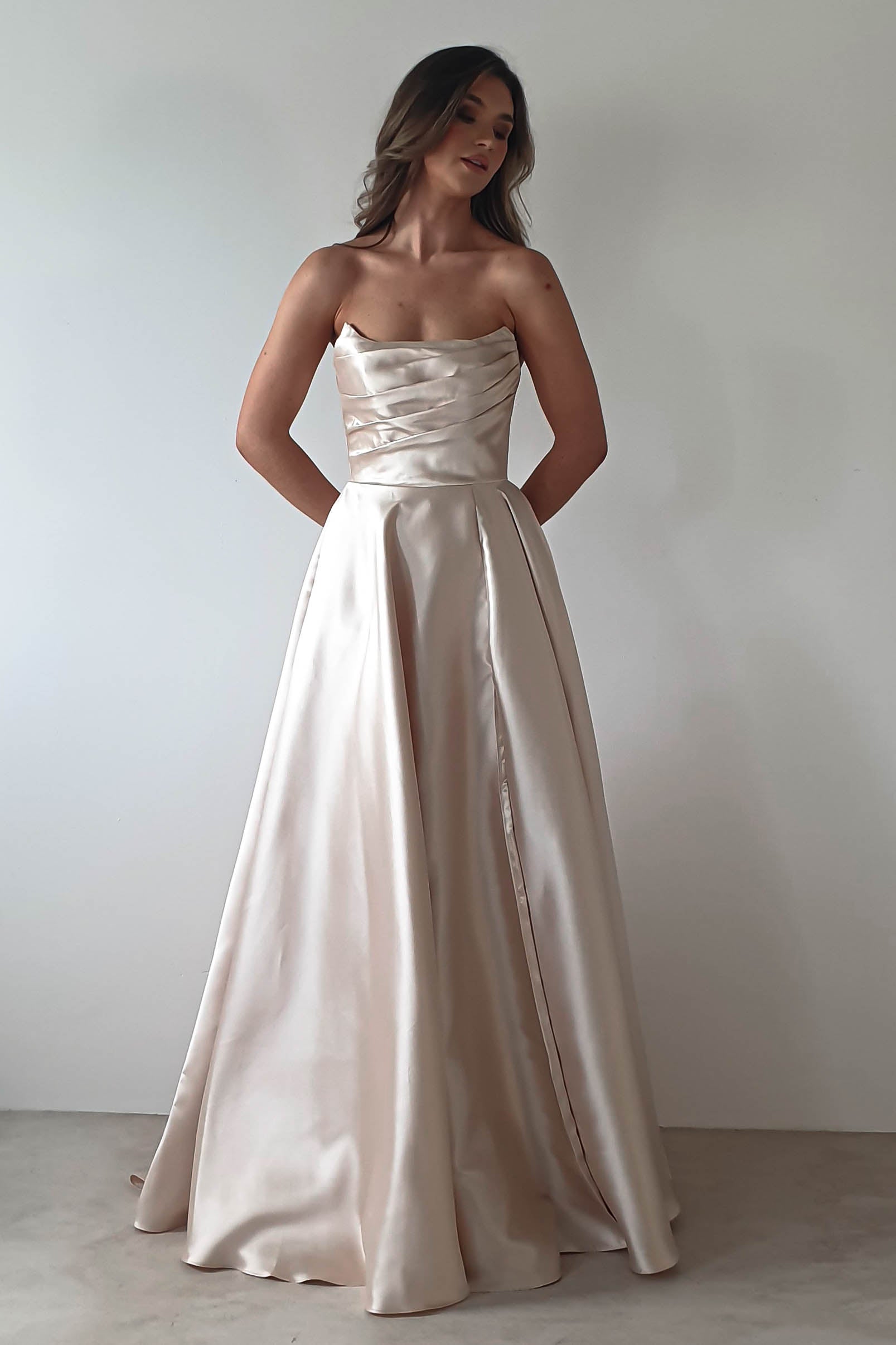A woman in a strapless satin gown stands against a plain white background, the dress featuring an elegant A-line skirt and fitted bodice. Her long, wavy hair cascades over her shoulders, and her hands rest in the gowns pockets, ready for the formal event ahead.