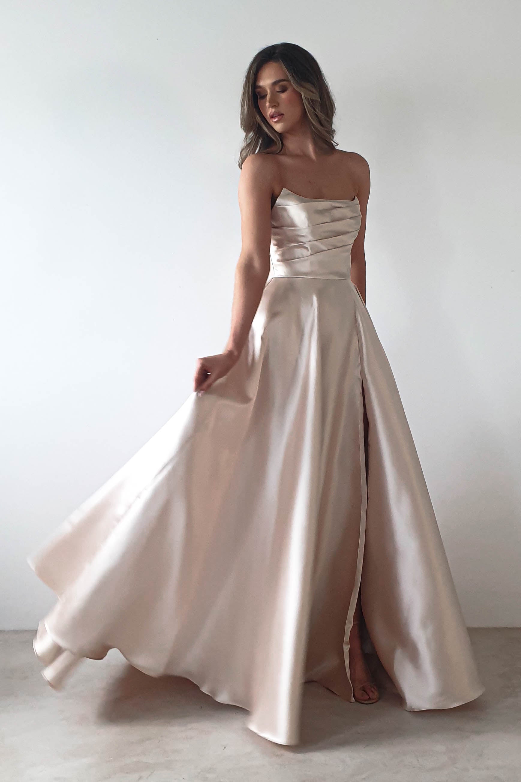 Woman in a strapless, satin beige gown poses gracefully against a plain white background. The formal event-ready dress boasts an A-line skirt with a flowing slit, and she has long, wavy brown hair cascading down her shoulders.